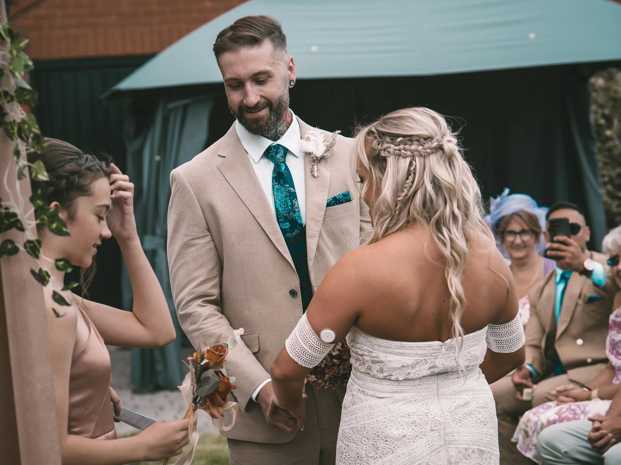 A man in a beige suit and a woman in a white dress exchange wedding vows outdoors, surrounded by guests and greenery.