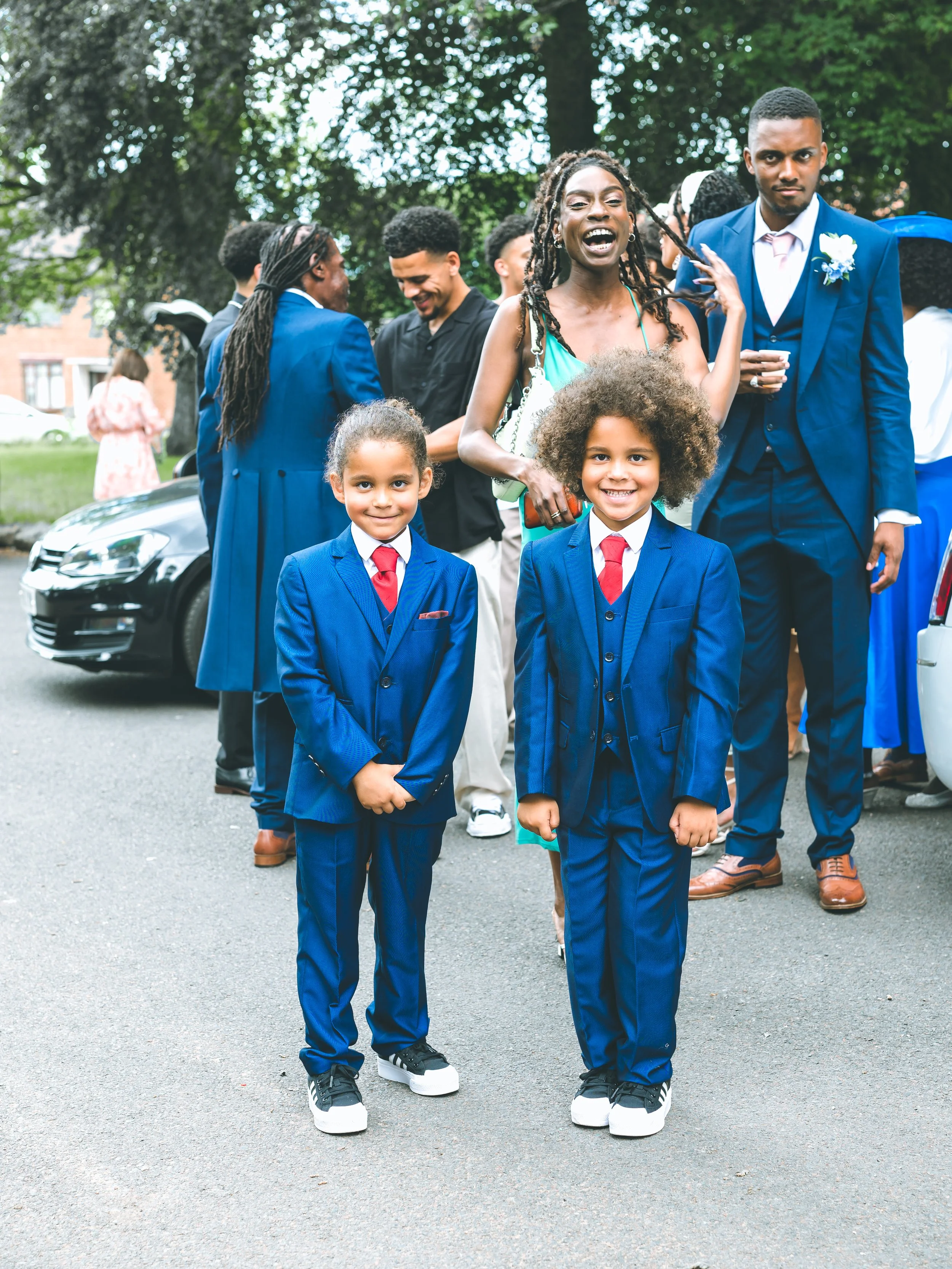 Two young boys in blue suits with red ties standing on a street, smiling at the camera. Behind them, a group of adults dressed in formal attire are socializing outdoors, with trees and a parking lot in the background.