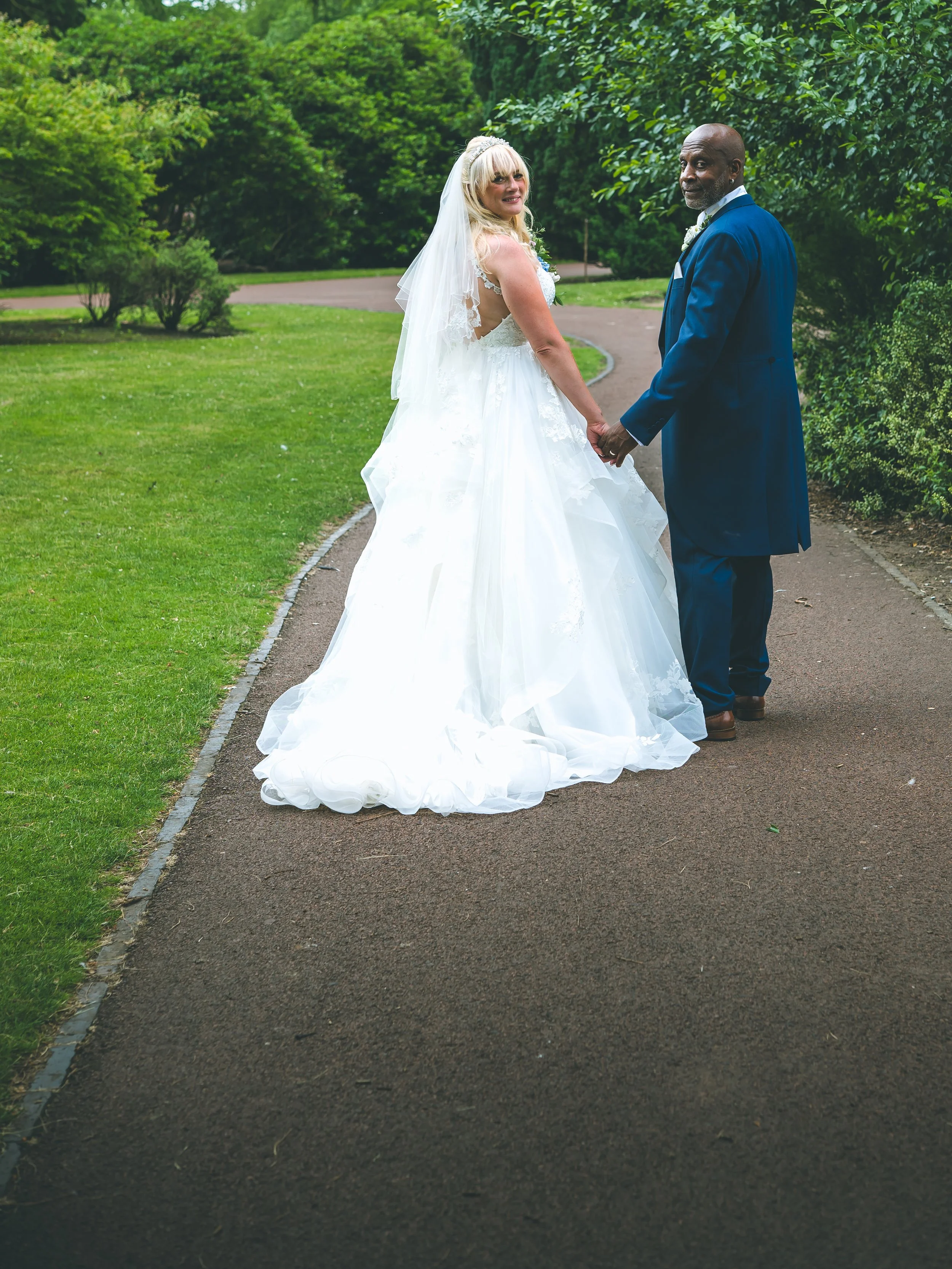 A bride and groom holding hands on a park pathway, the bride in a white wedding dress and veil, the groom in a blue suit, surrounded by green trees.