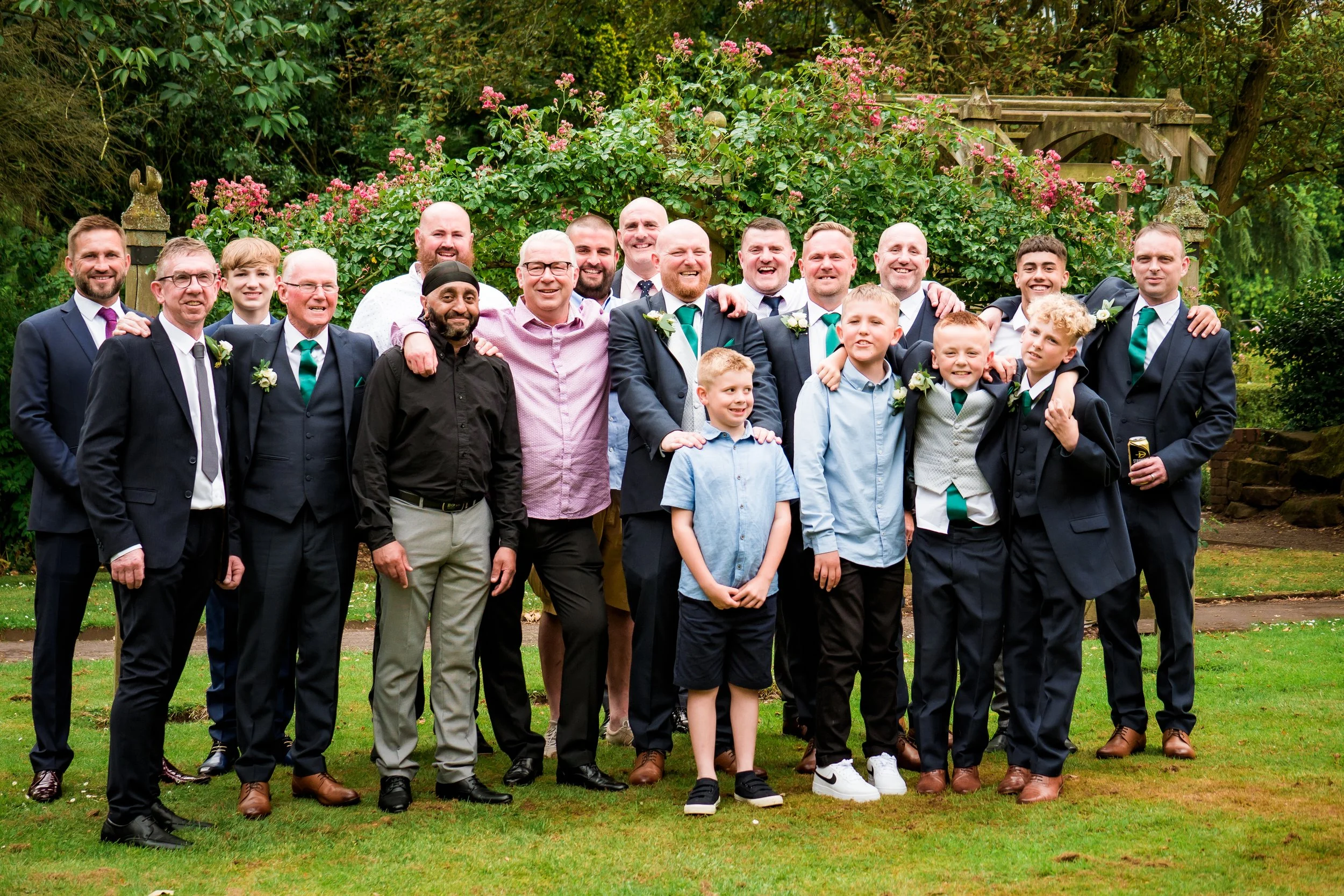 A group of men and boys dressed in formal attire, smiling and posing outdoors in front of green bushes and pink flowers.