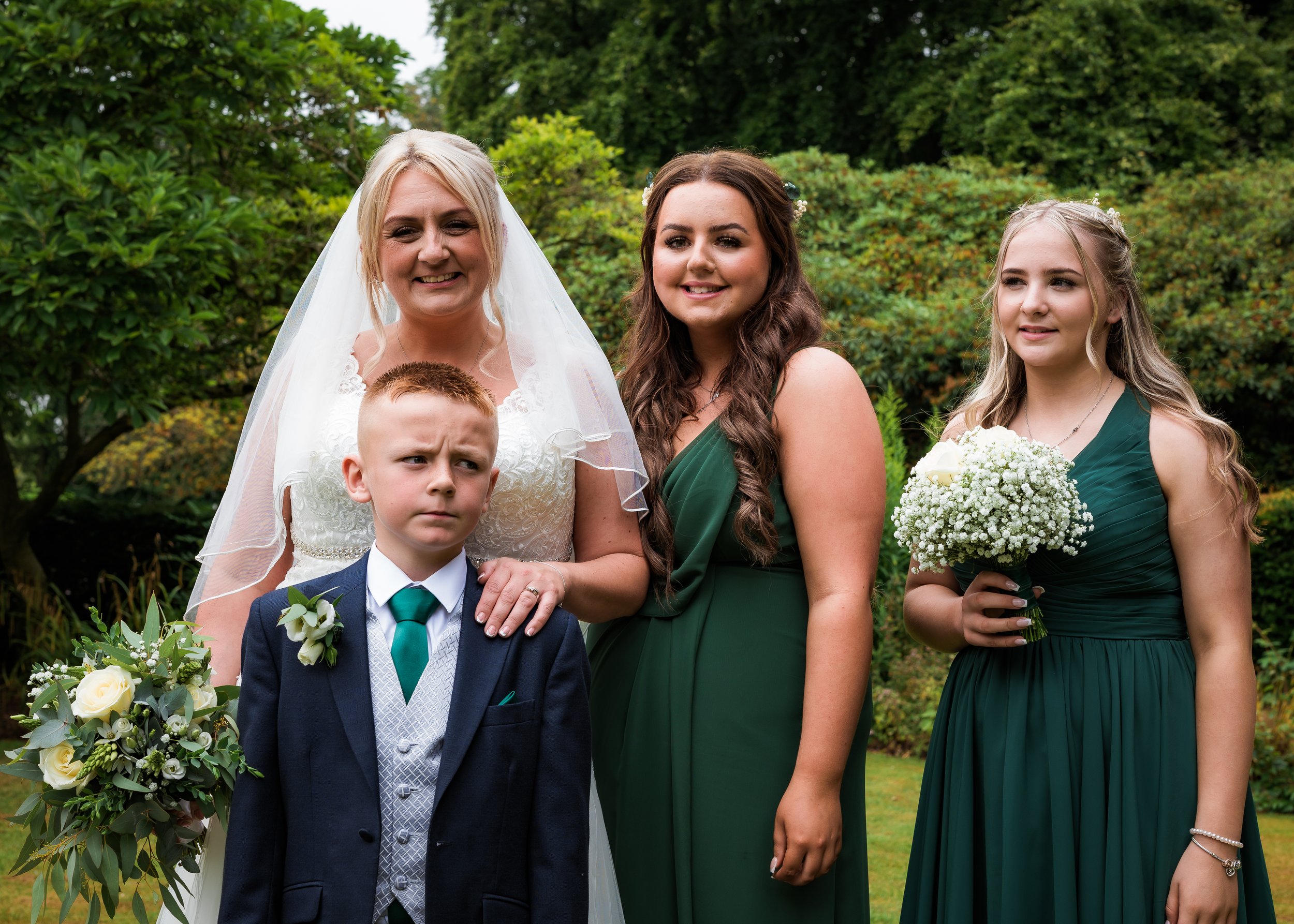 A group of four people including a woman in a wedding dress, a young boy in a suit, a woman in a green dress, and a young girl in a green dress holding a bouquet, standing outdoors with green trees in the background.