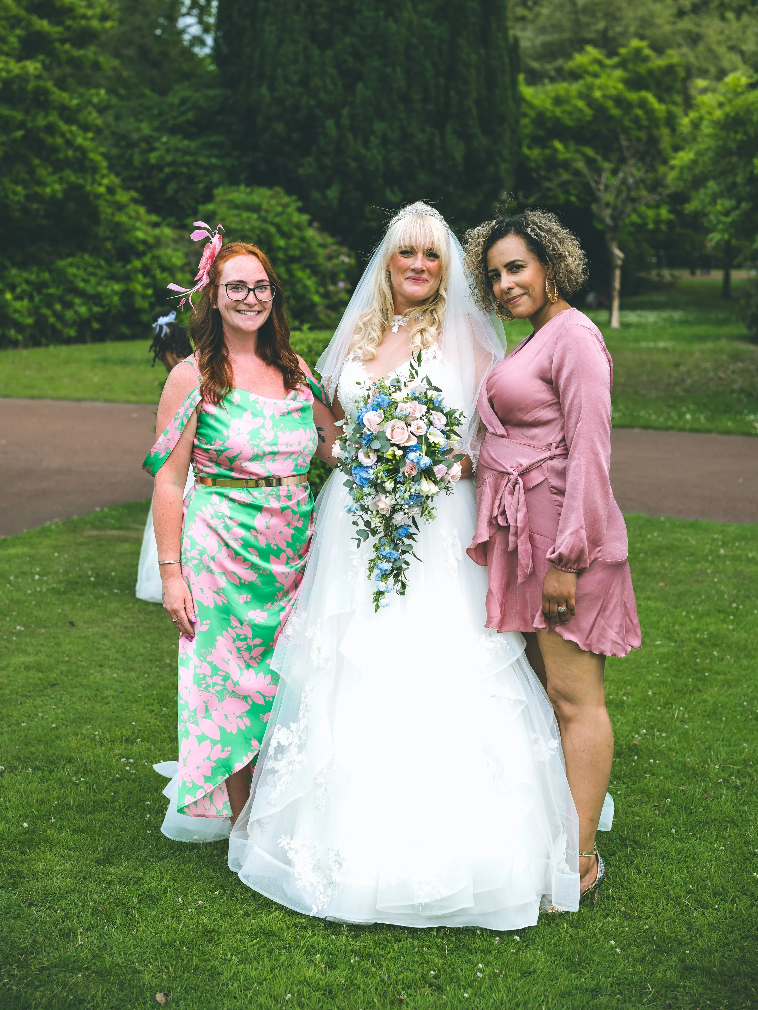 Three women standing on a grassy area with trees in the background. The woman in the middle is wearing a white wedding dress and veil, holding a bouquet of flowers. The other two women are dressed in colorful outfits, one in a pink and green dress wi