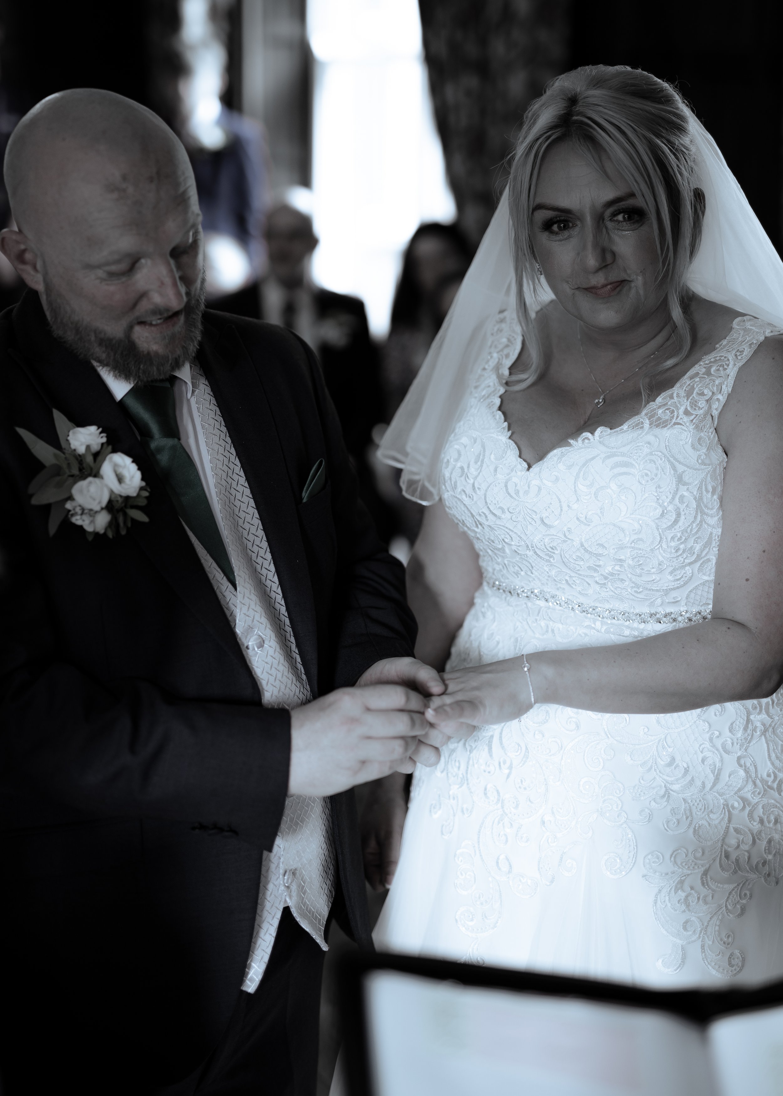 A bride and groom exchanging vows, holding hands during their wedding ceremony. The bride is wearing a white lace wedding dress and veil, and the groom is dressed in a dark suit with a boutonniere. They are standing inside a rustic venue with exposed