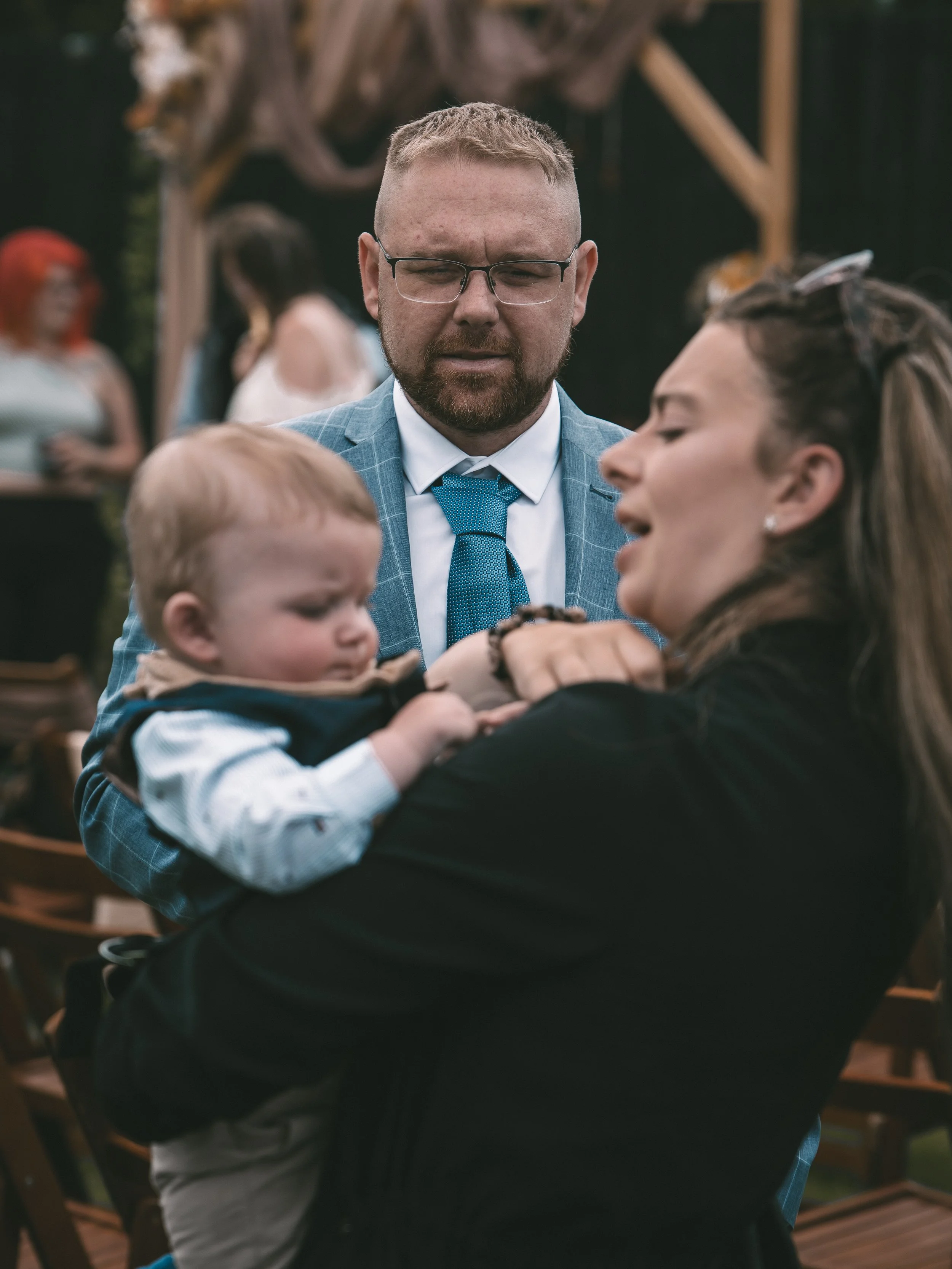 A man with glasses in a suit and blue tie is holding a young boy, and they are talking to a woman with long hair. There are other people and greenery in the blurred background.