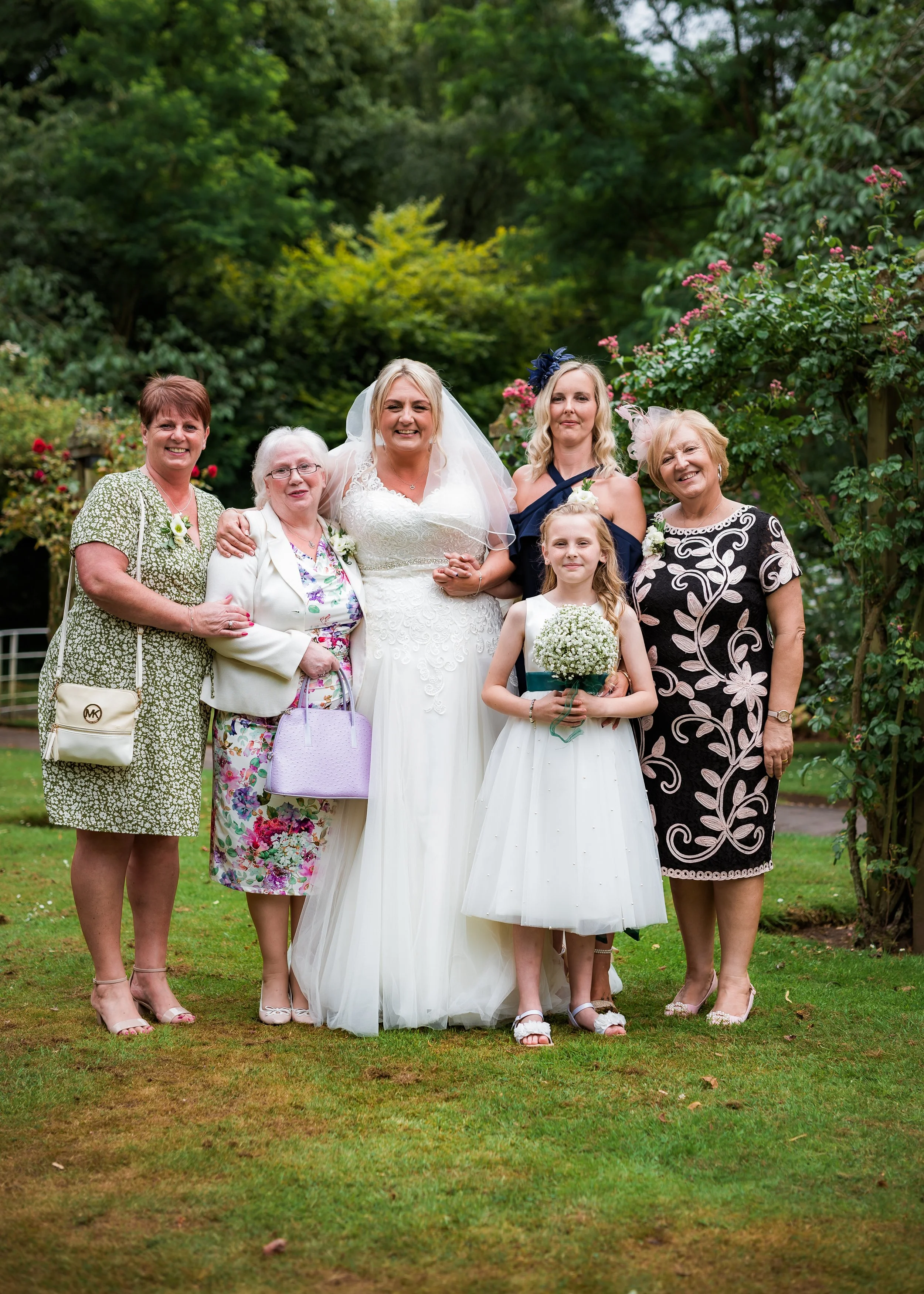 A group of six women and one young girl posing outdoors on a grassy area, with trees and bushes in the background. The woman in the white wedding dress is surrounded by the other women, all dressed in various dresses. The young girl is holding a bouq