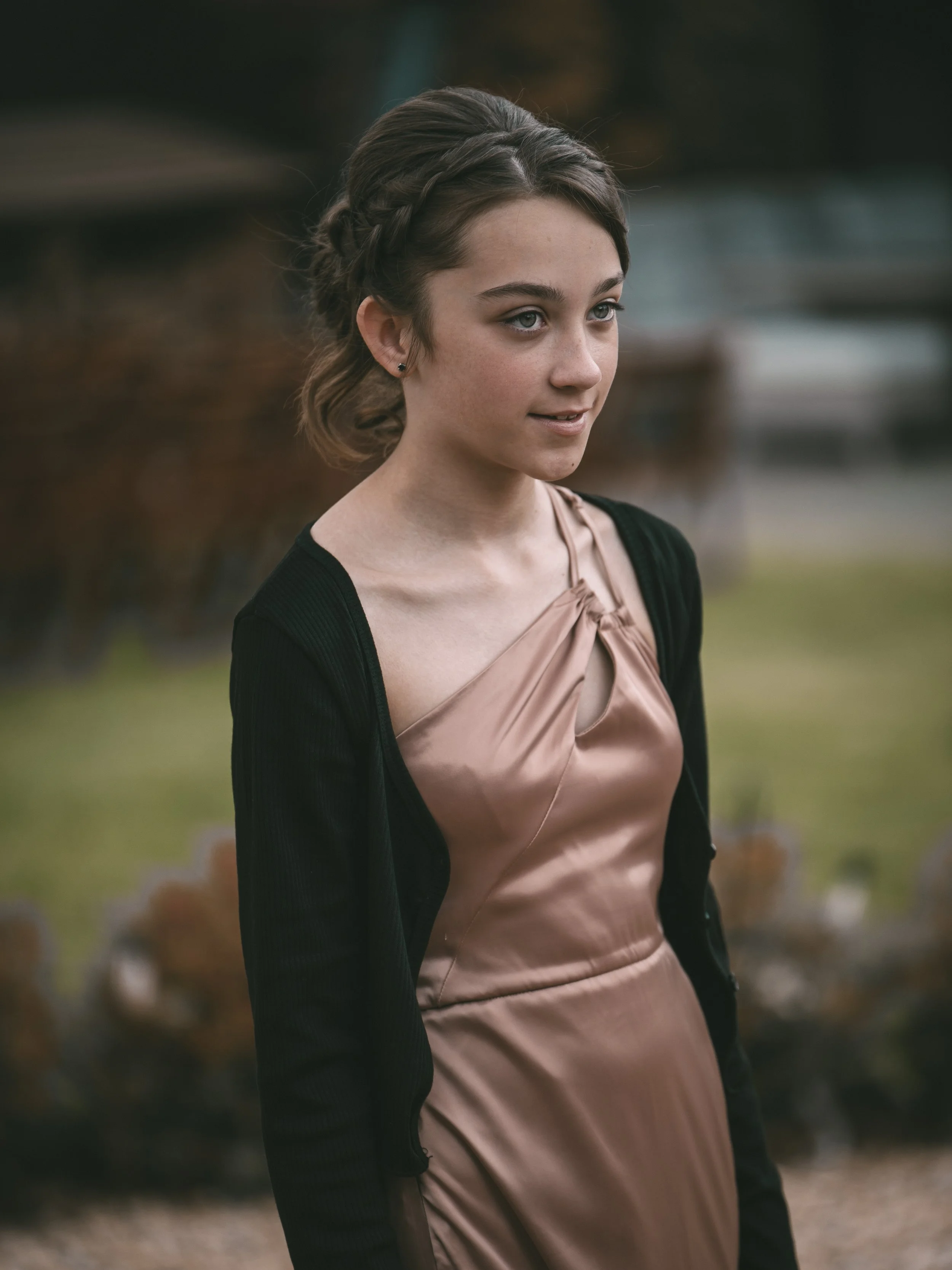 A young woman with braided brown hair and light skin, wearing a beige satin dress and black cardigan, standing outdoors with blurred trees in the background.