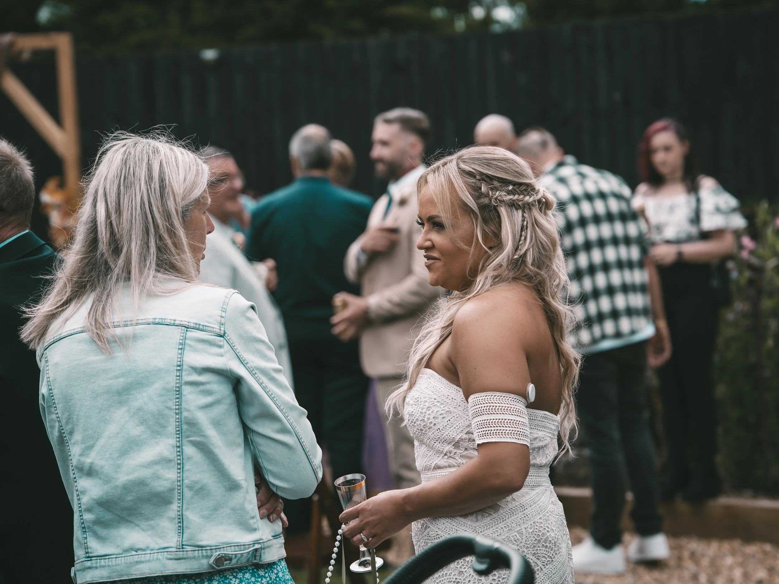 People socializing outdoors during a celebration, with a woman in a white dress holding a glass of champagne and a rosary, engaging in conversation with a woman in a denim jacket, while others chat in the background.