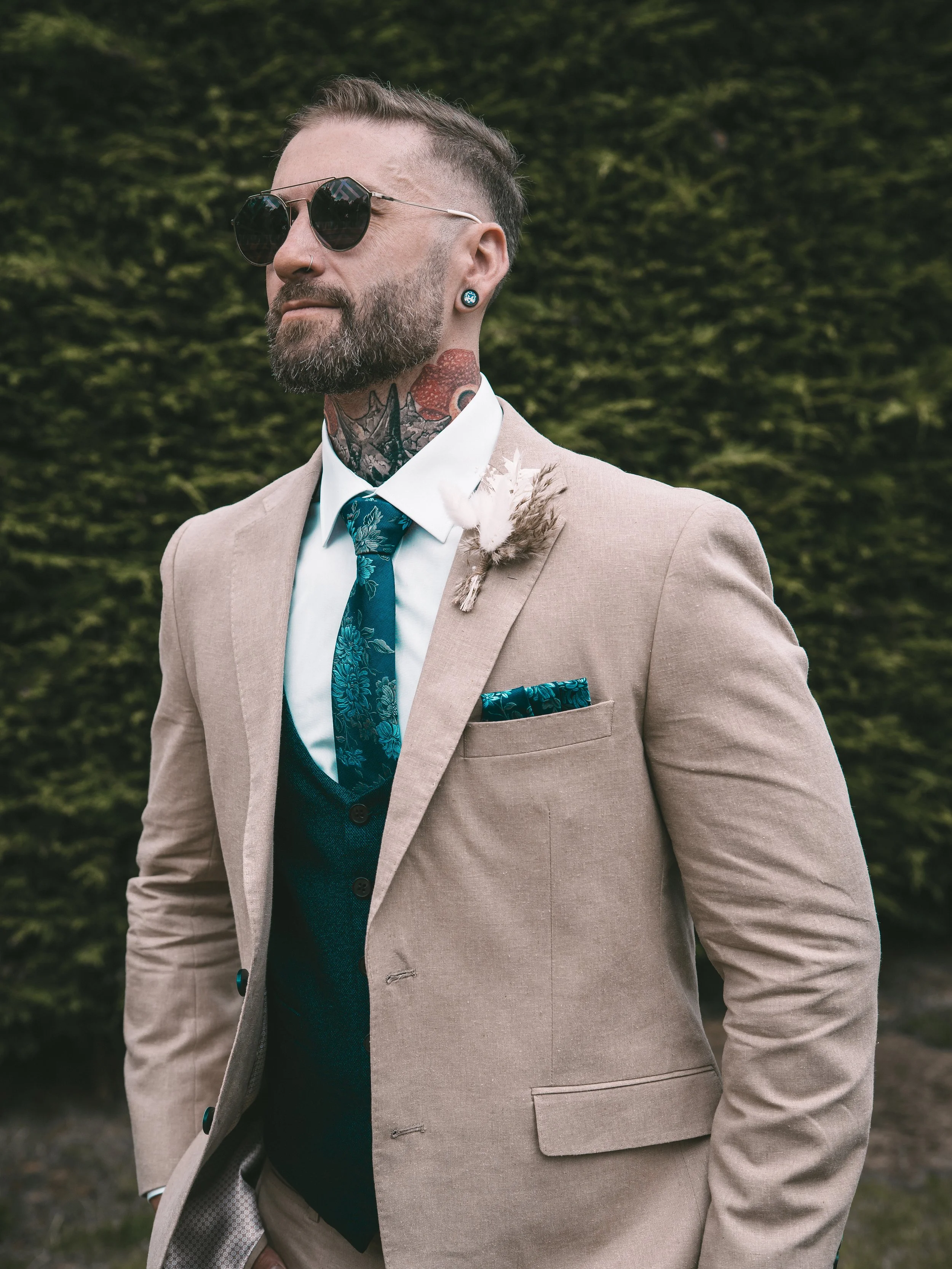 A man wearing sunglasses, a beige suit, and a white shirt with a blue tie, standing outdoors in front of greenery.