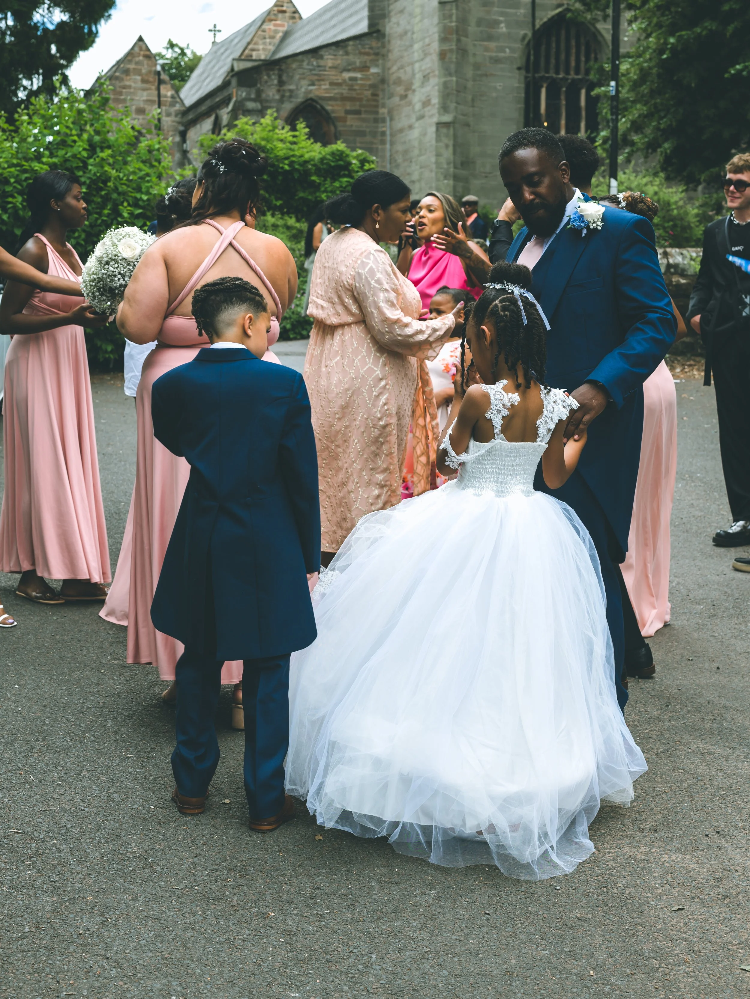A group of people dressed in formal attire, including a young girl in a white dress and a man in a blue suit, gathered outdoors near a historic building with stone walls and a cross on the roof, during a wedding celebration.