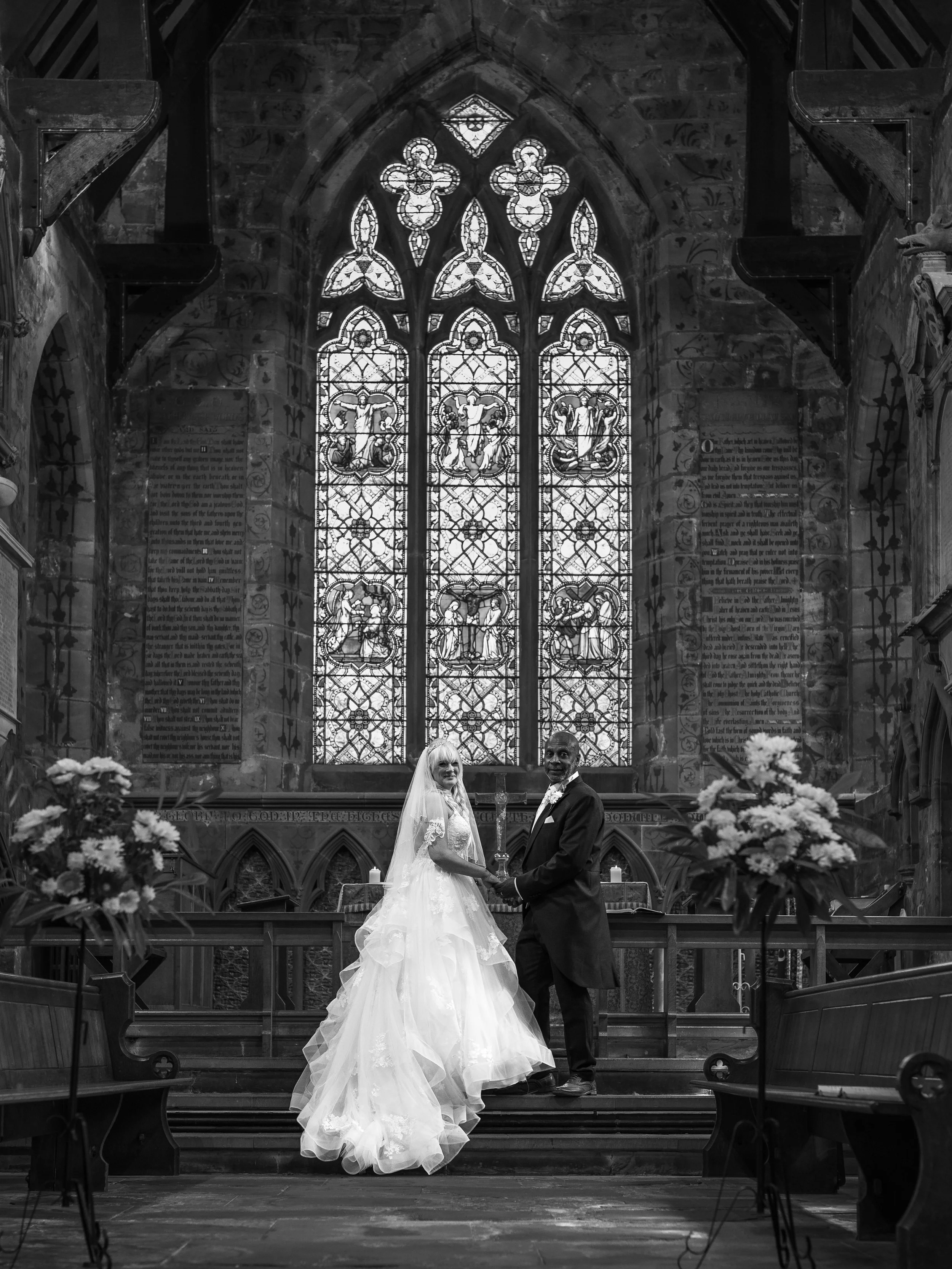 Black and white photo of a bride and groom standing hand in hand at the altar inside a church with high stained glass windows. The church has wooden pews, floral arrangements, and religious wall plaques.
