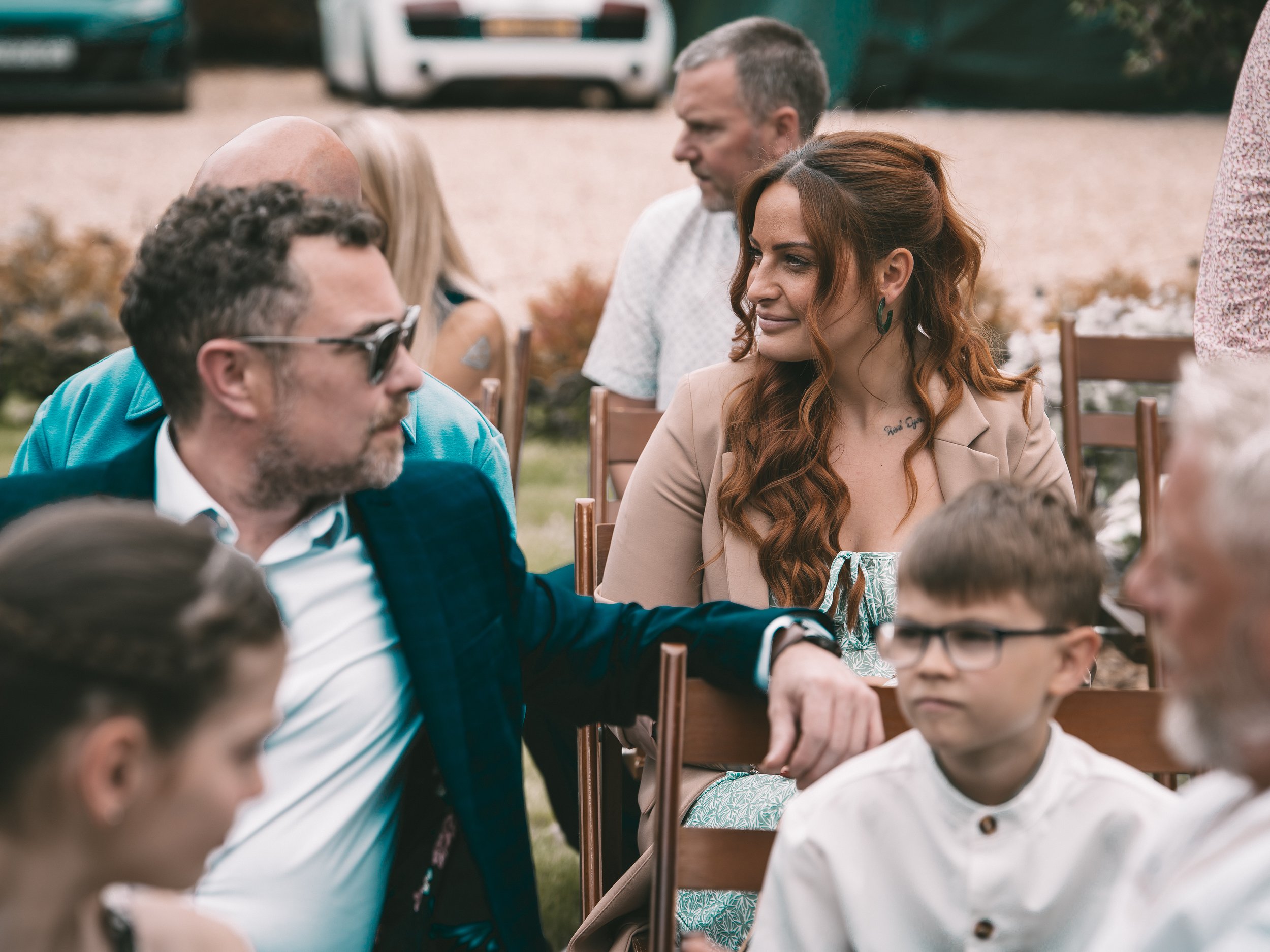 Group of people sitting outdoors at a gathering, engaging in conversation. There are adults and children, with cars in the background.
