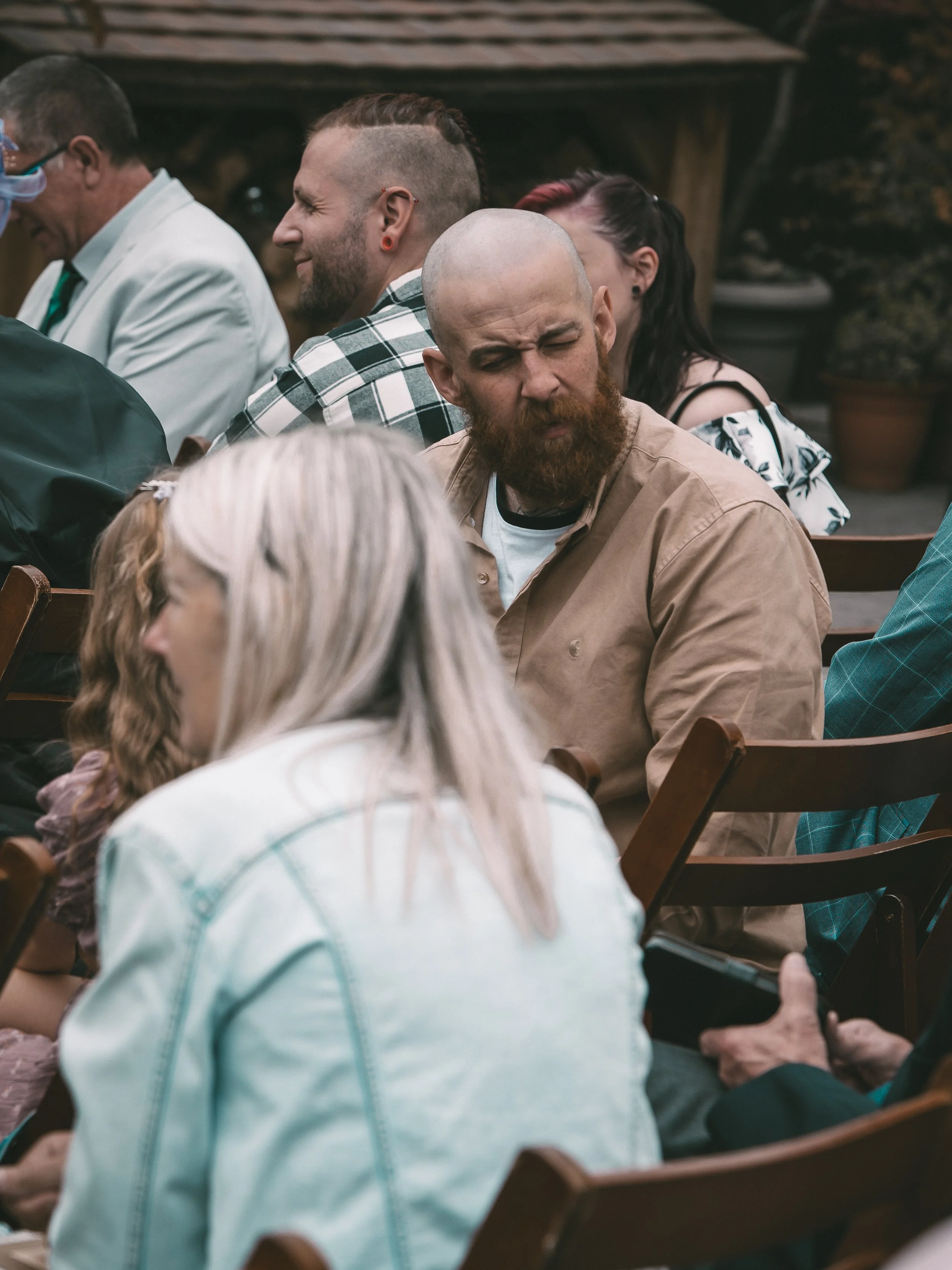 Group of diverse people sitting at outdoor event, some engaged in conversations, some looking down or to the side, with wooden seating and a potted plant in the background.