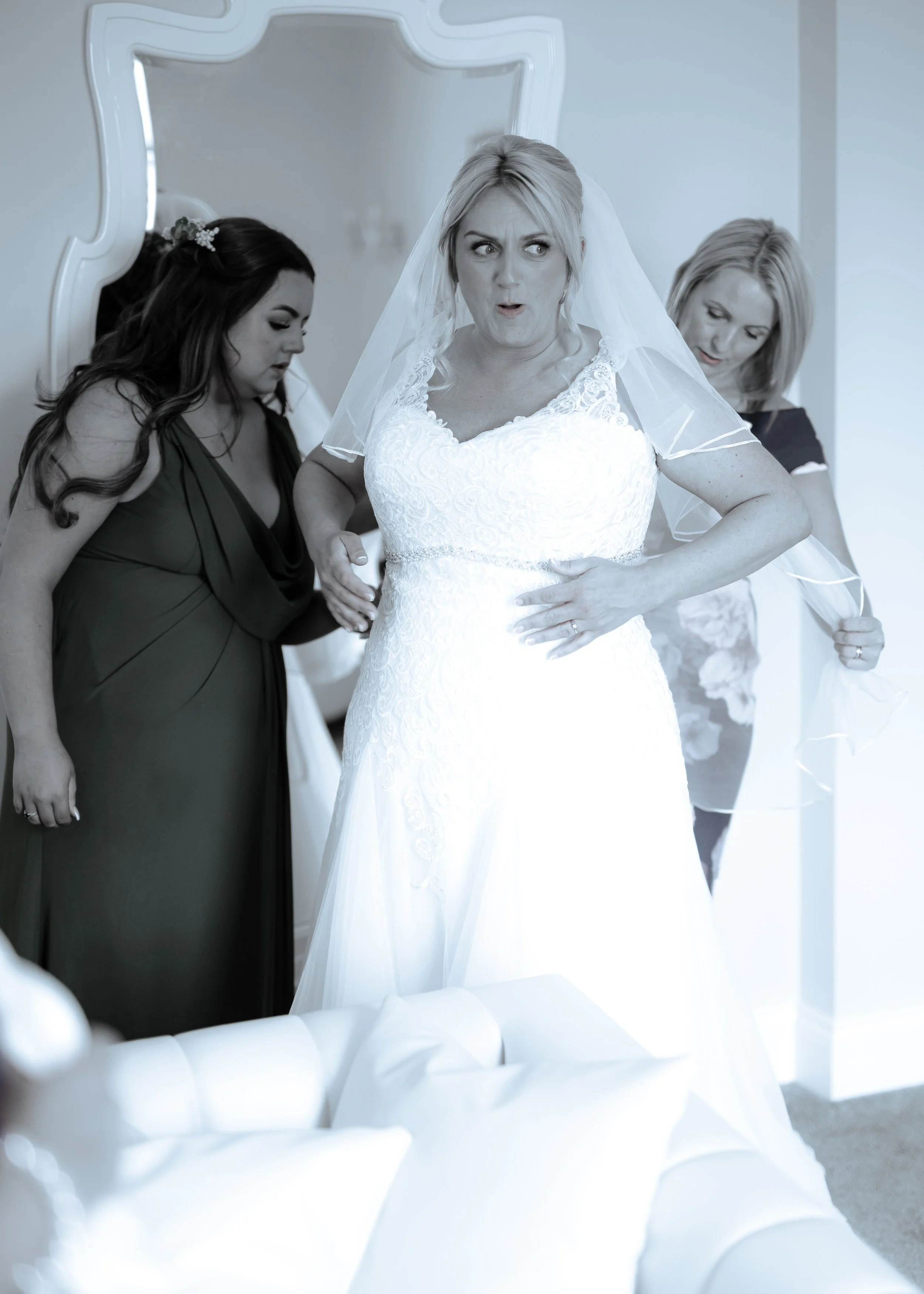 A bride in a wedding dress with a veil is surrounded by women helping her prepare, with one woman touching her dress and another adjusting her veil.