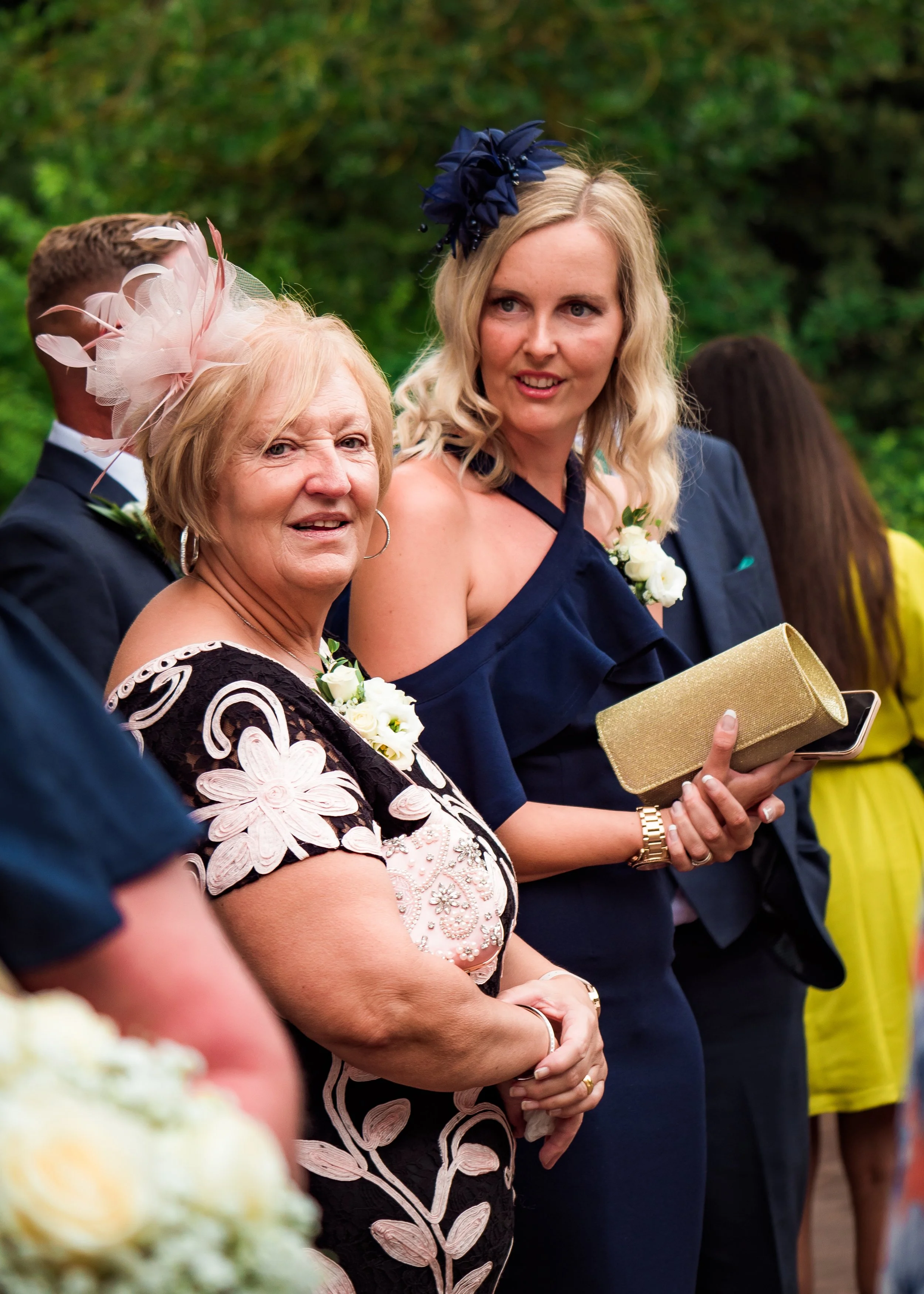 Group of people dressed in formal attire attending a outdoor event, with women wearing floral dresses and men in suits, some women with decorative headpieces.