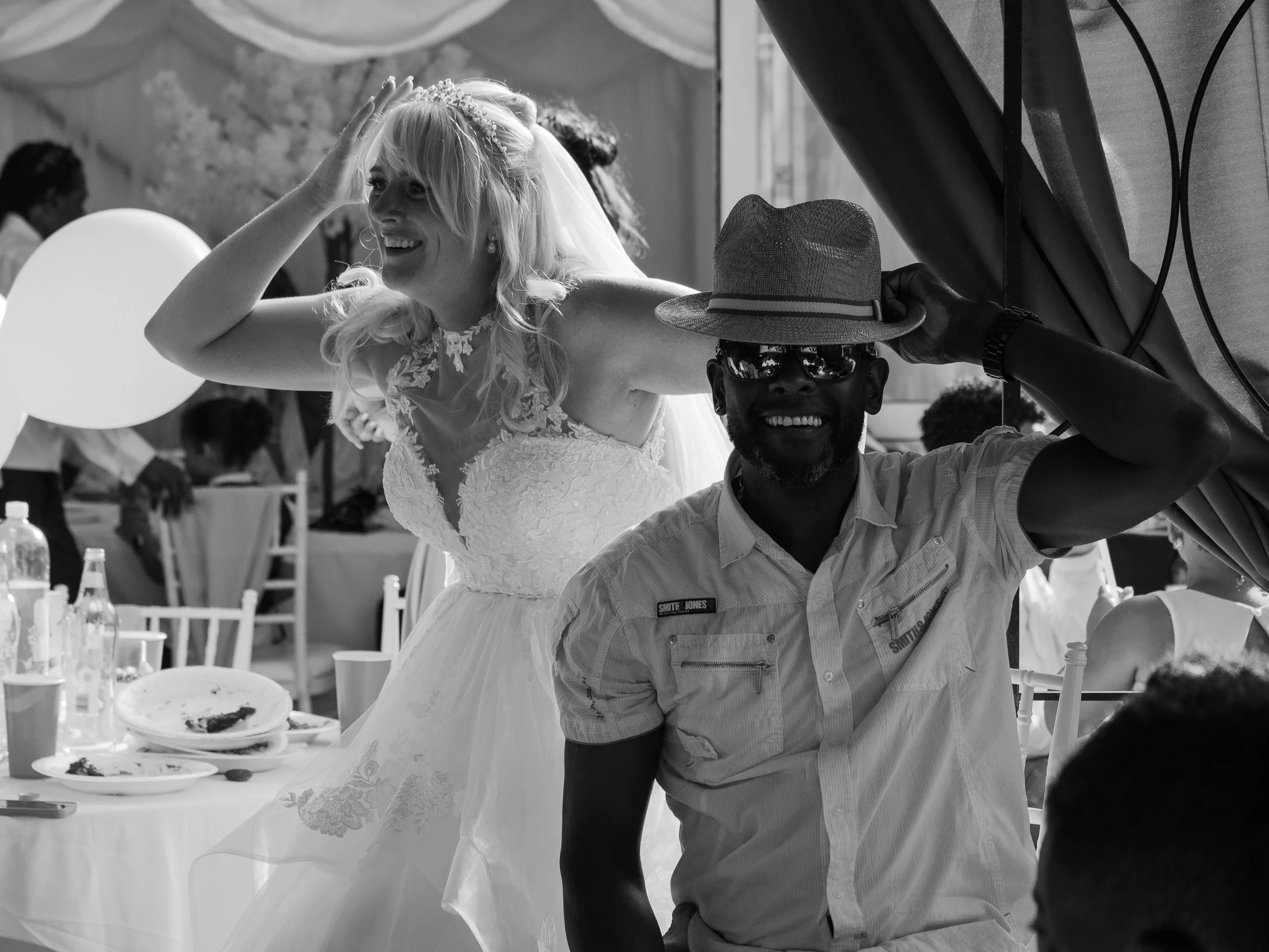 A bride with long blonde hair wearing a wedding dress and a tiara, smiling and gesturing with her hand while seated at a celebration, with a man wearing sunglasses and a hat beside her, and people and tables visible in the background.