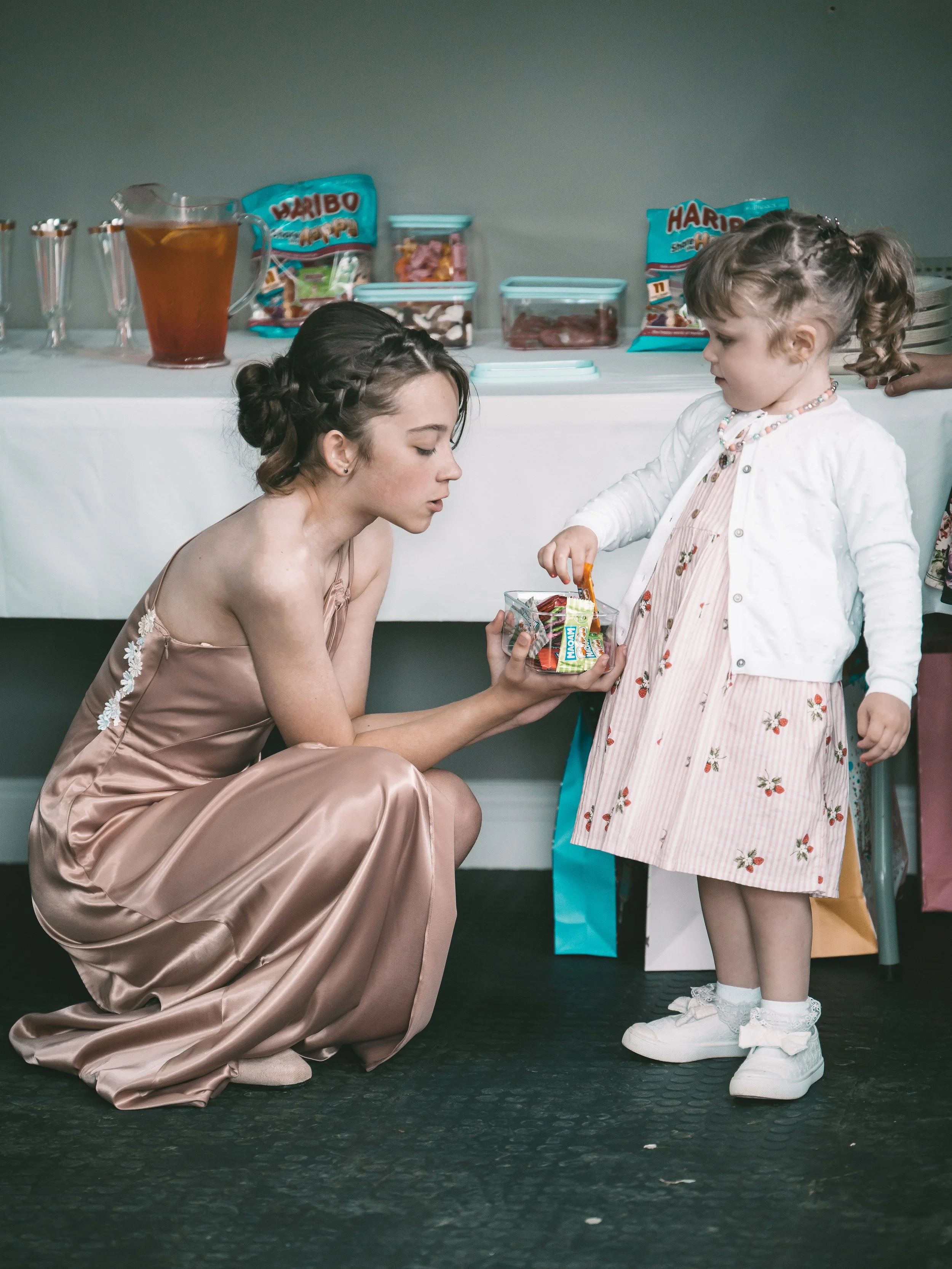 A woman crouches on the floor handing a bag of candy to a young girl in a pink floral dress. The girl stands with her hand extended towards the woman, wearing a beaded necklace, a white cardigan, a pink striped dress, and white sneakers. In the backg