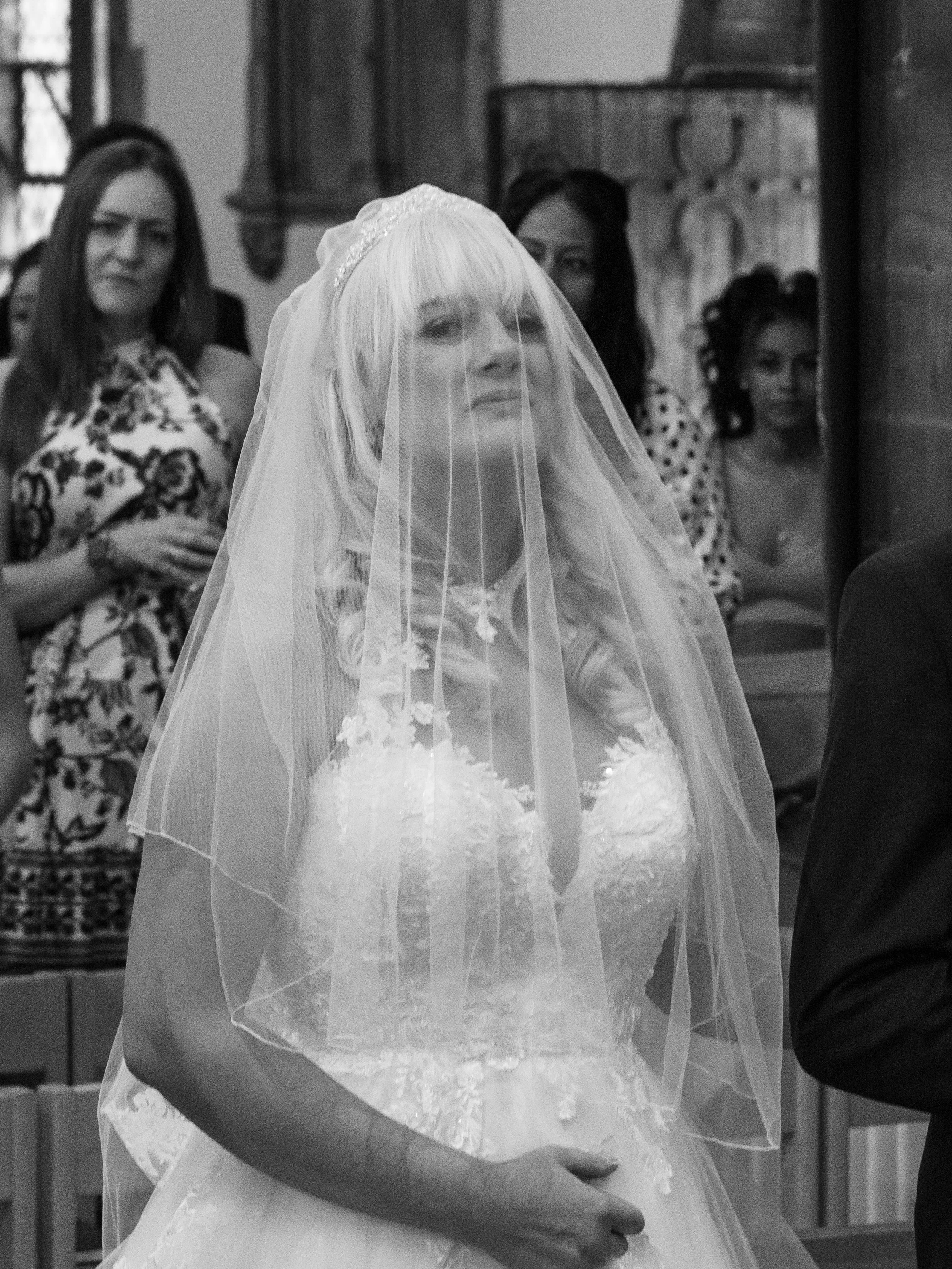 A bride wearing a veil and wedding dress standing with her arm around someone, with three women in the background indoors.