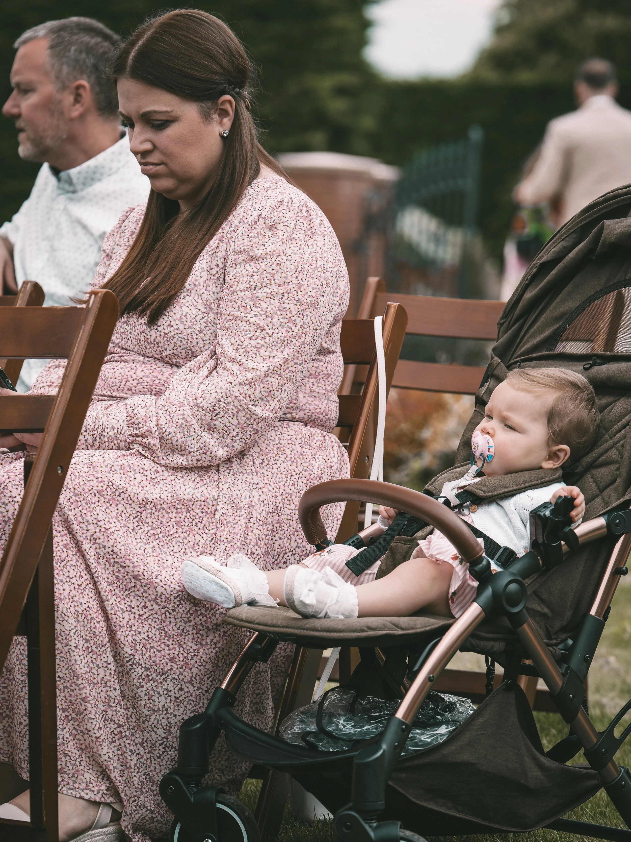 A woman sitting outdoors on a wooden bench, looking down, with a man next to her and a young girl in a stroller nearby. The scene appears to be in a park or garden with blurred greenery in the background.