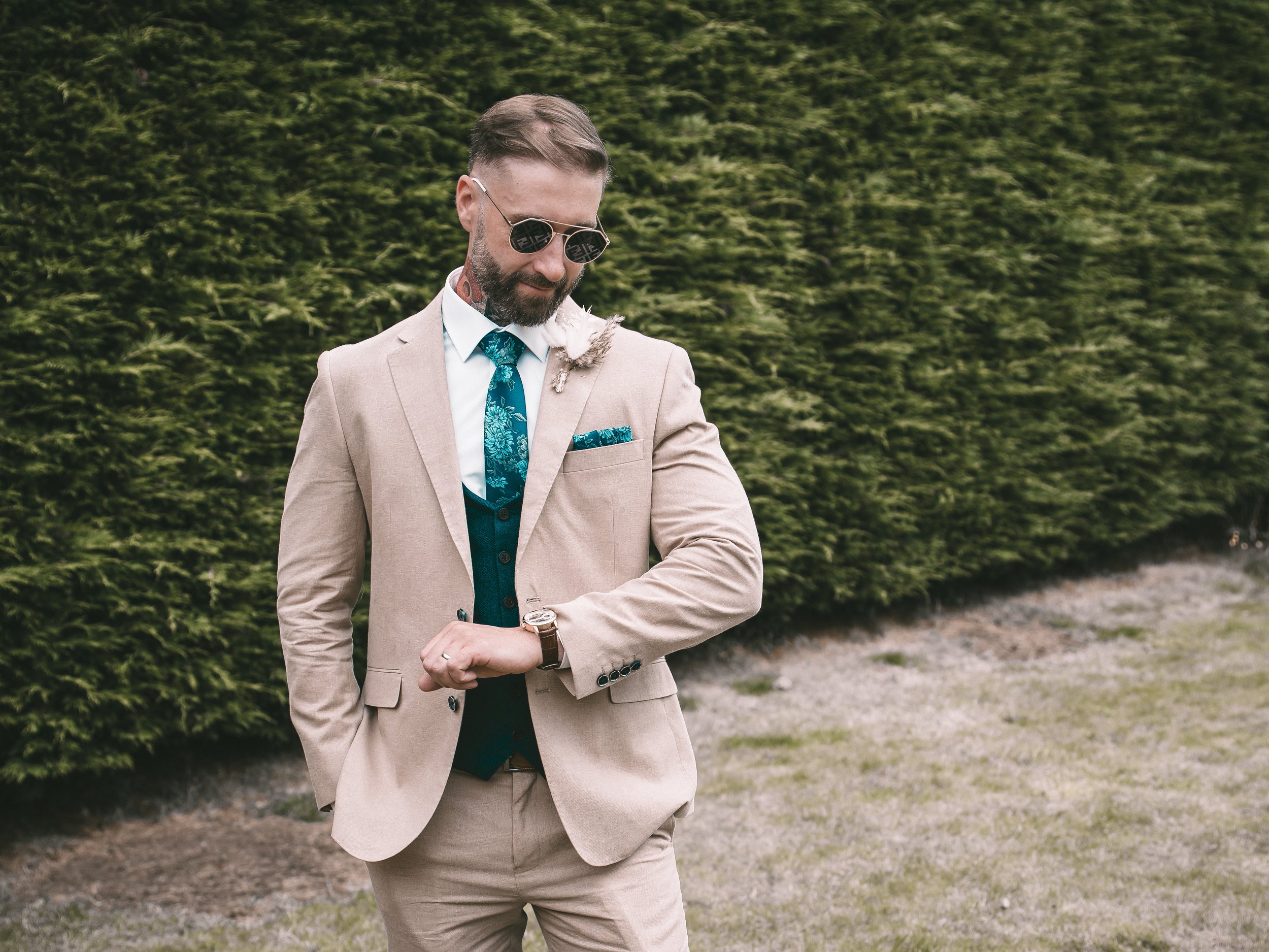 A man in a beige suit with sunglasses, a blue patterned tie, and a watch, standing outdoors in front of green bushes.
