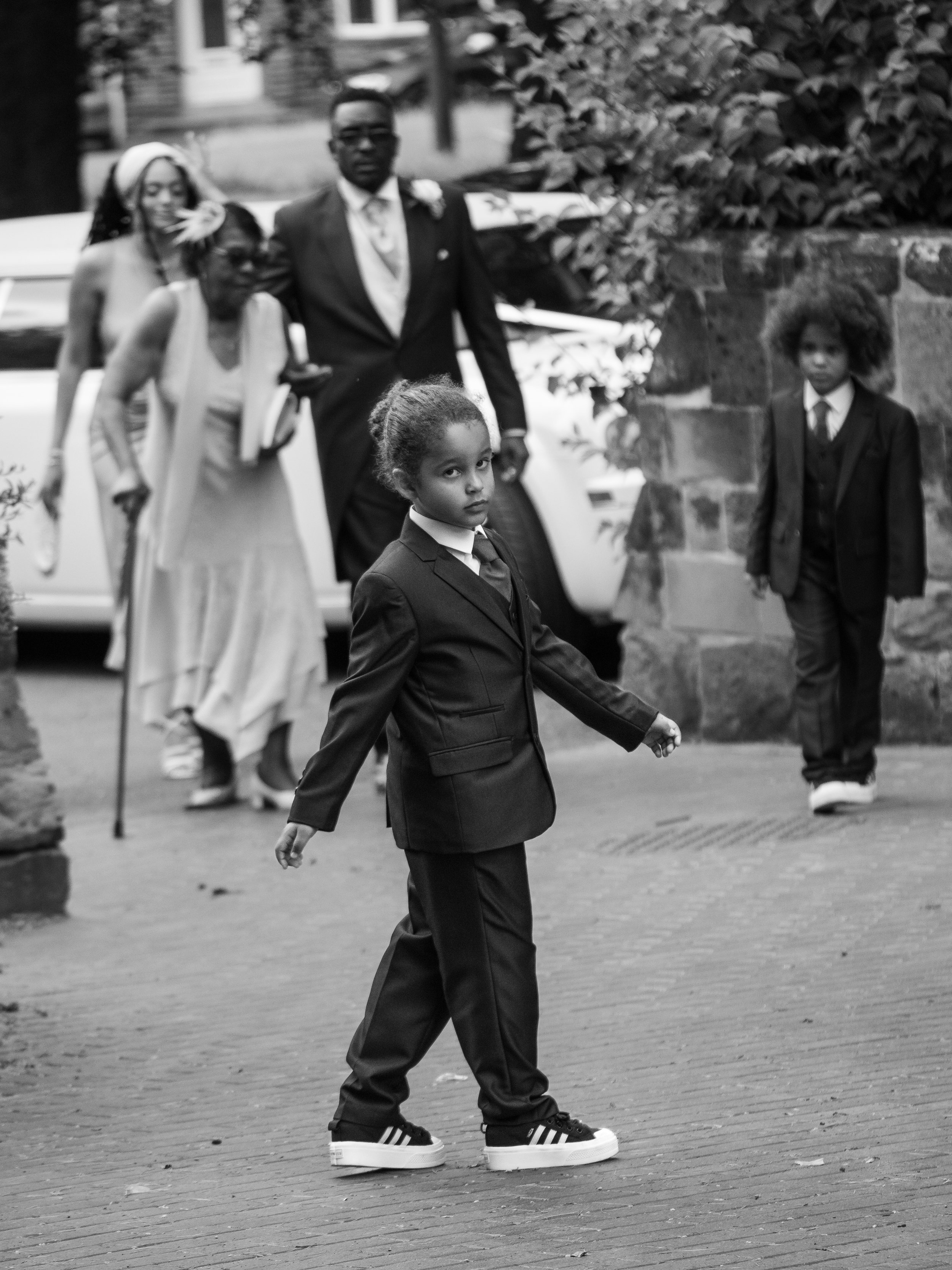 A group of people, including children and adults, gathered outdoors on a street, with some dressed in formal clothing, and one person wearing oversized sneakers.