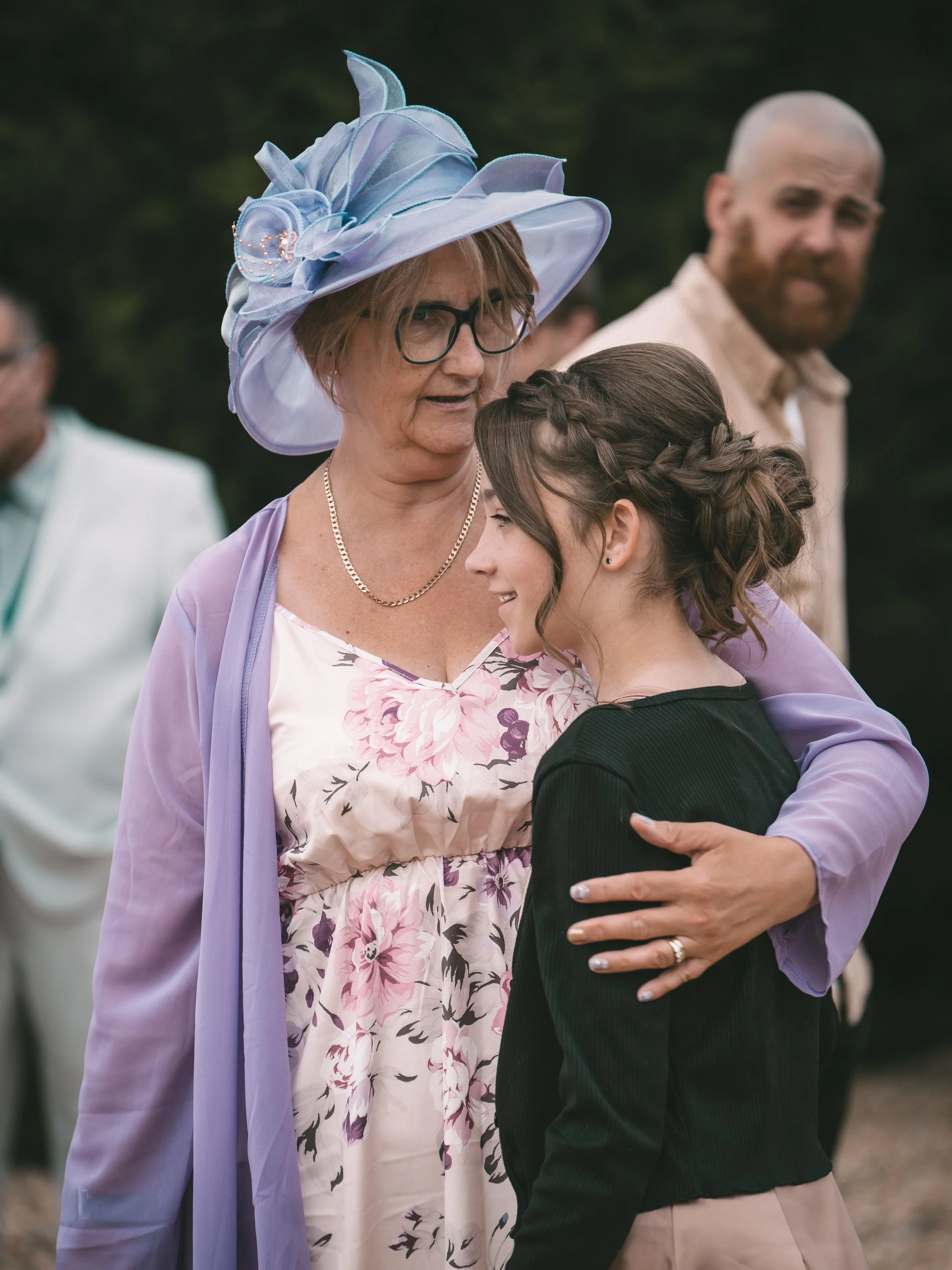 An elderly woman wearing a large blue hat with floral details and a lavender shawl, hugging a young woman with long braid and dark clothing at an outdoor gathering.