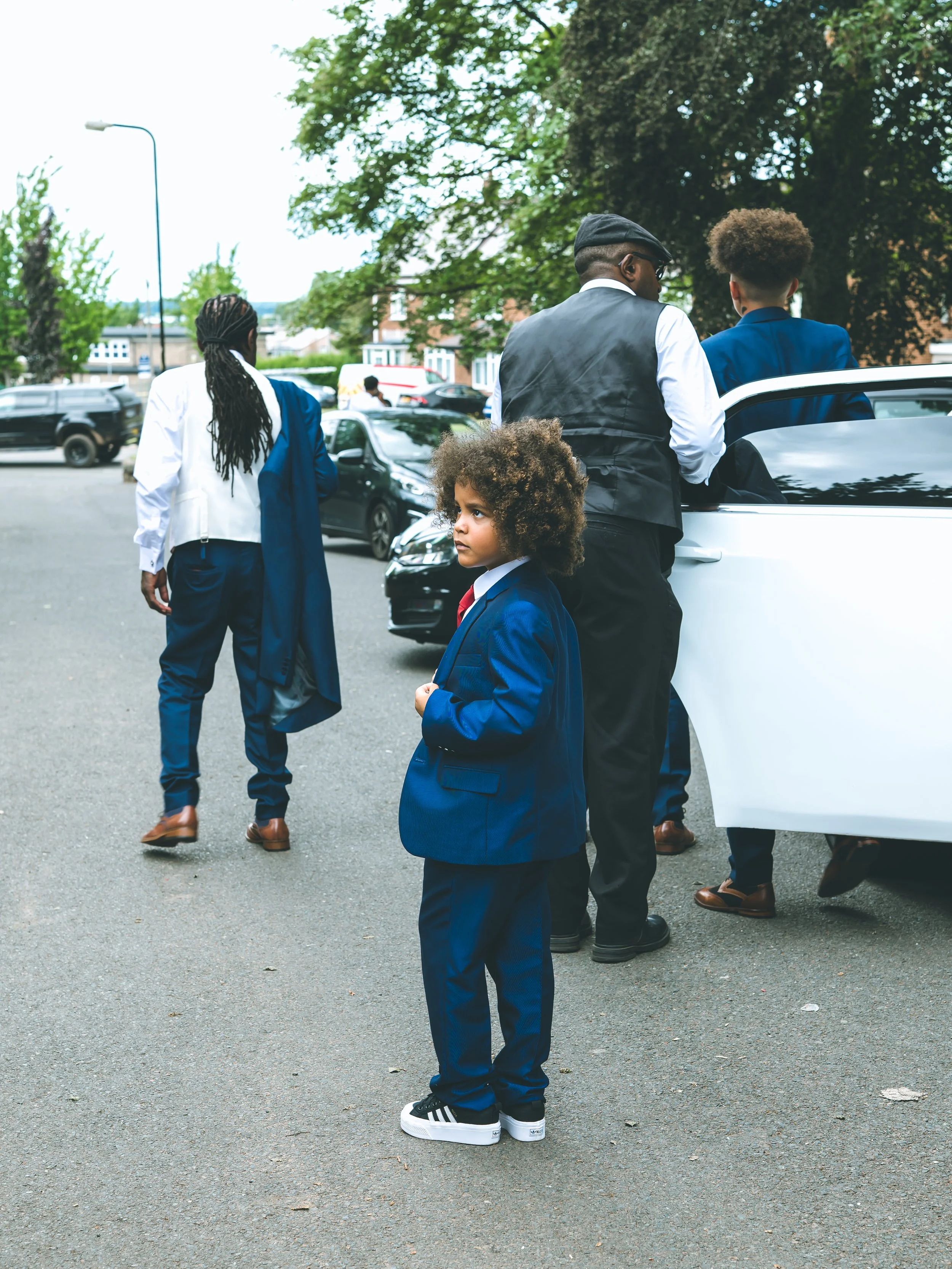 A group of four people, including a young boy in a blue suit, standing and talking outside near a white car on an overcast day with trees and parked cars in the background.