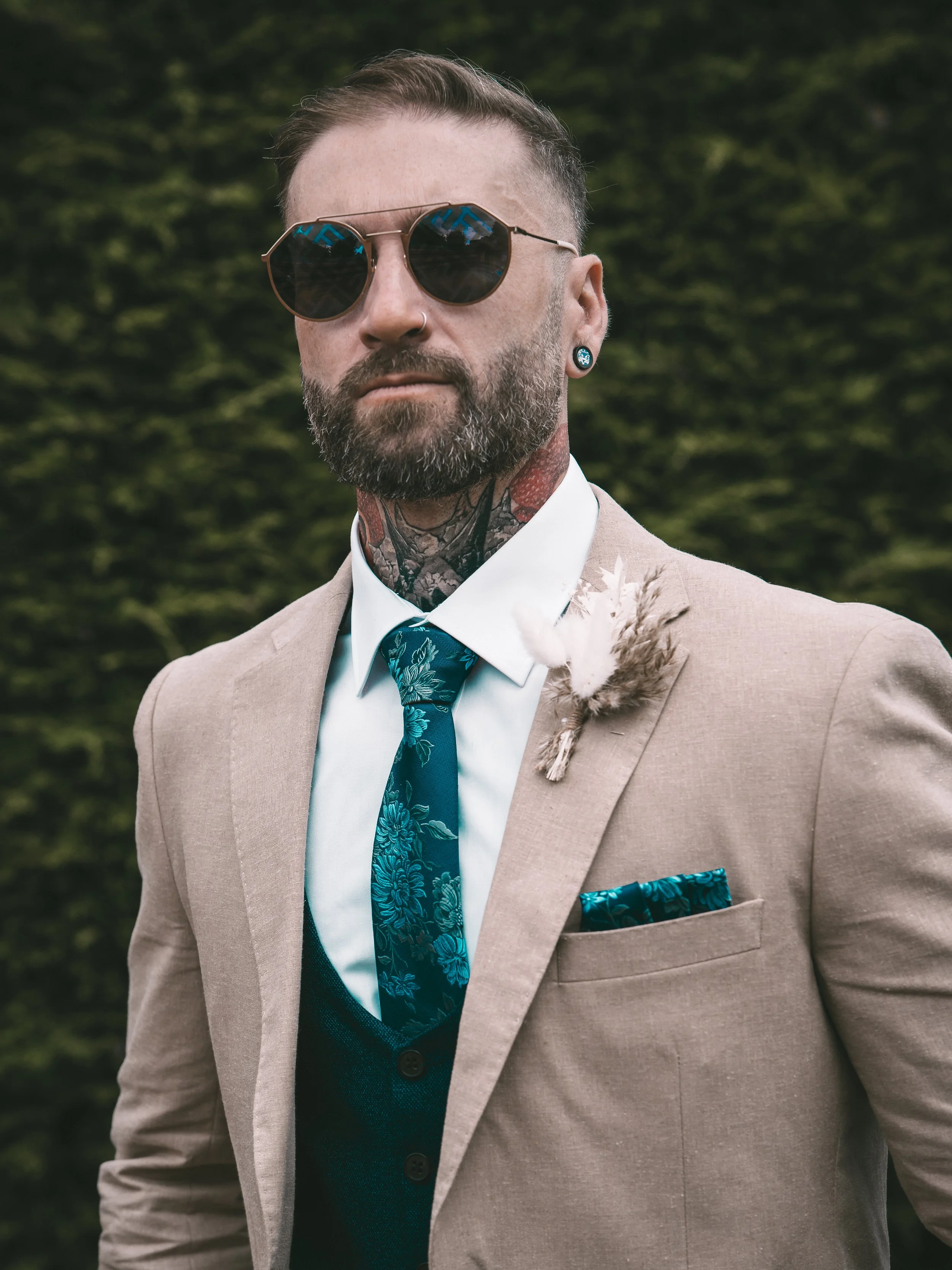 A man with a beard and tattoos wearing sunglasses, a tan suit, a floral tie, a white shirt, and a pocket square, standing outdoors with green trees in the background.