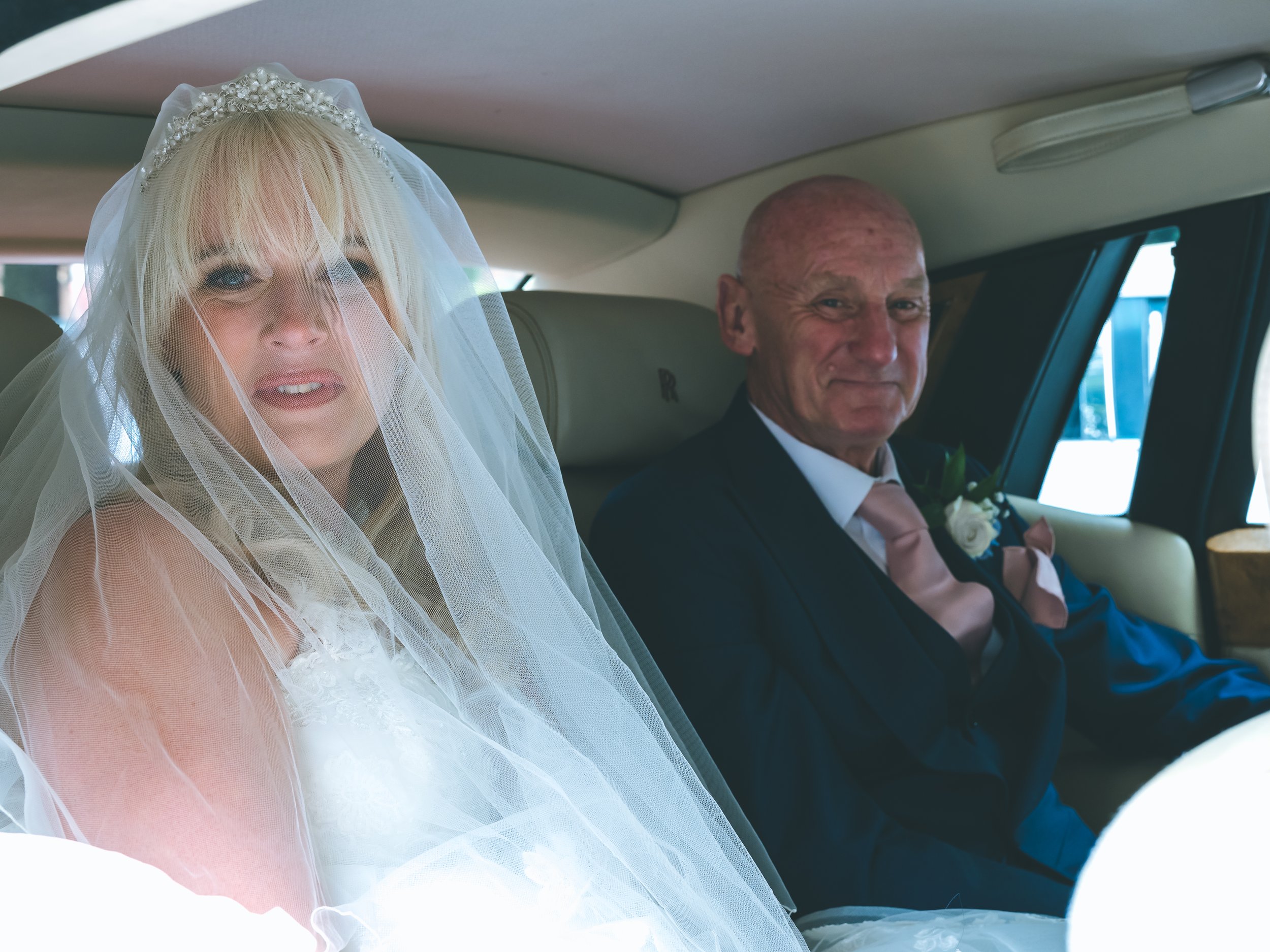 A bride with blonde hair wearing a wedding veil and tiara, sitting next to a groom in a car, both smiling.