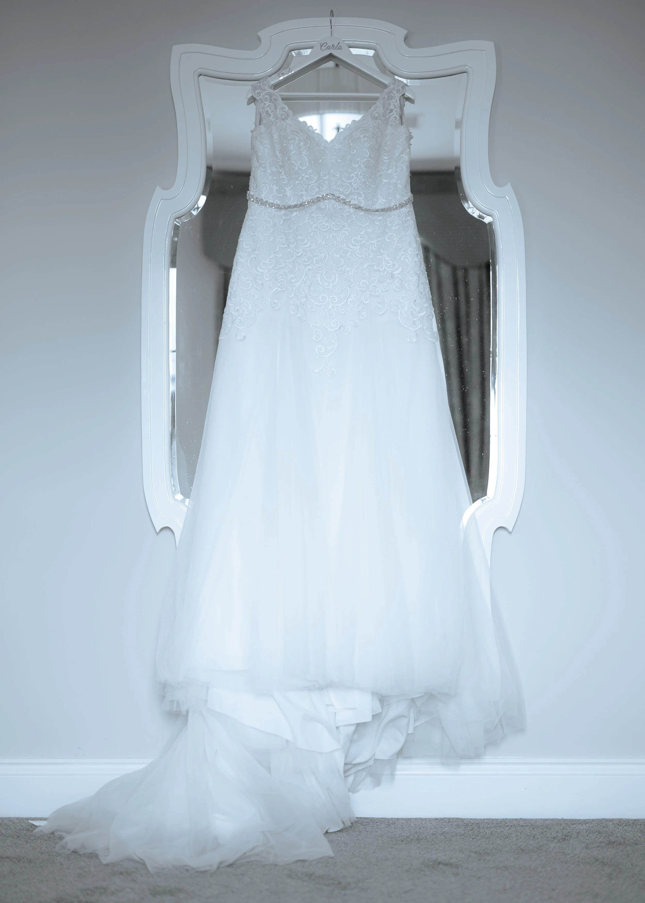 A white wedding dress hanging on a hanger with the name 'Carla' on it, reflected in a decorative mirror, with the train of the dress spilling onto the floor.