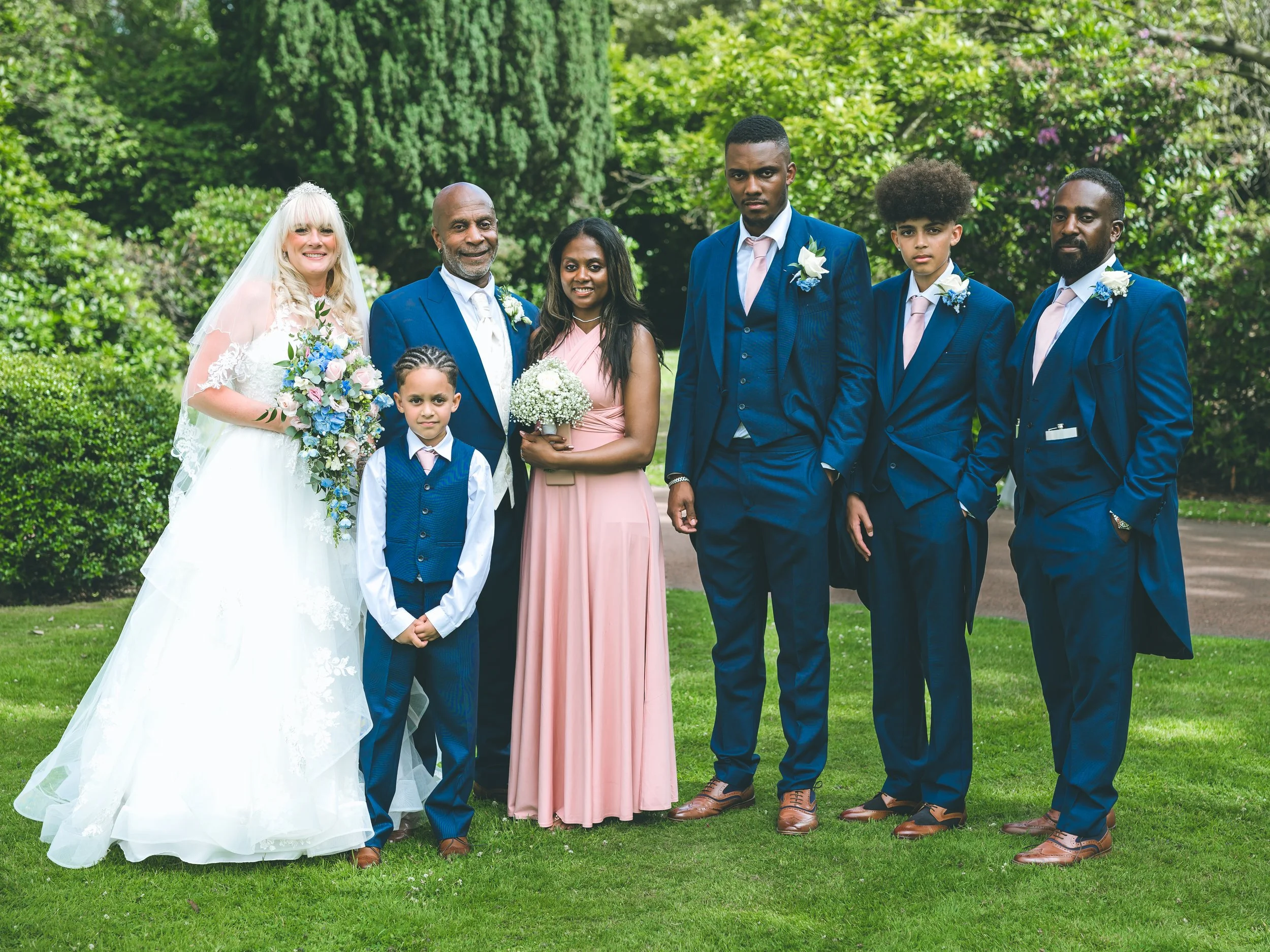 Group of wedding guests dressed in formal attire, standing outdoors in a lush green garden. One woman is in a white wedding gown holding a colorful bouquet, and a man is wearing a blue suit, white shirt, and tie. The group appears to be posing for a 