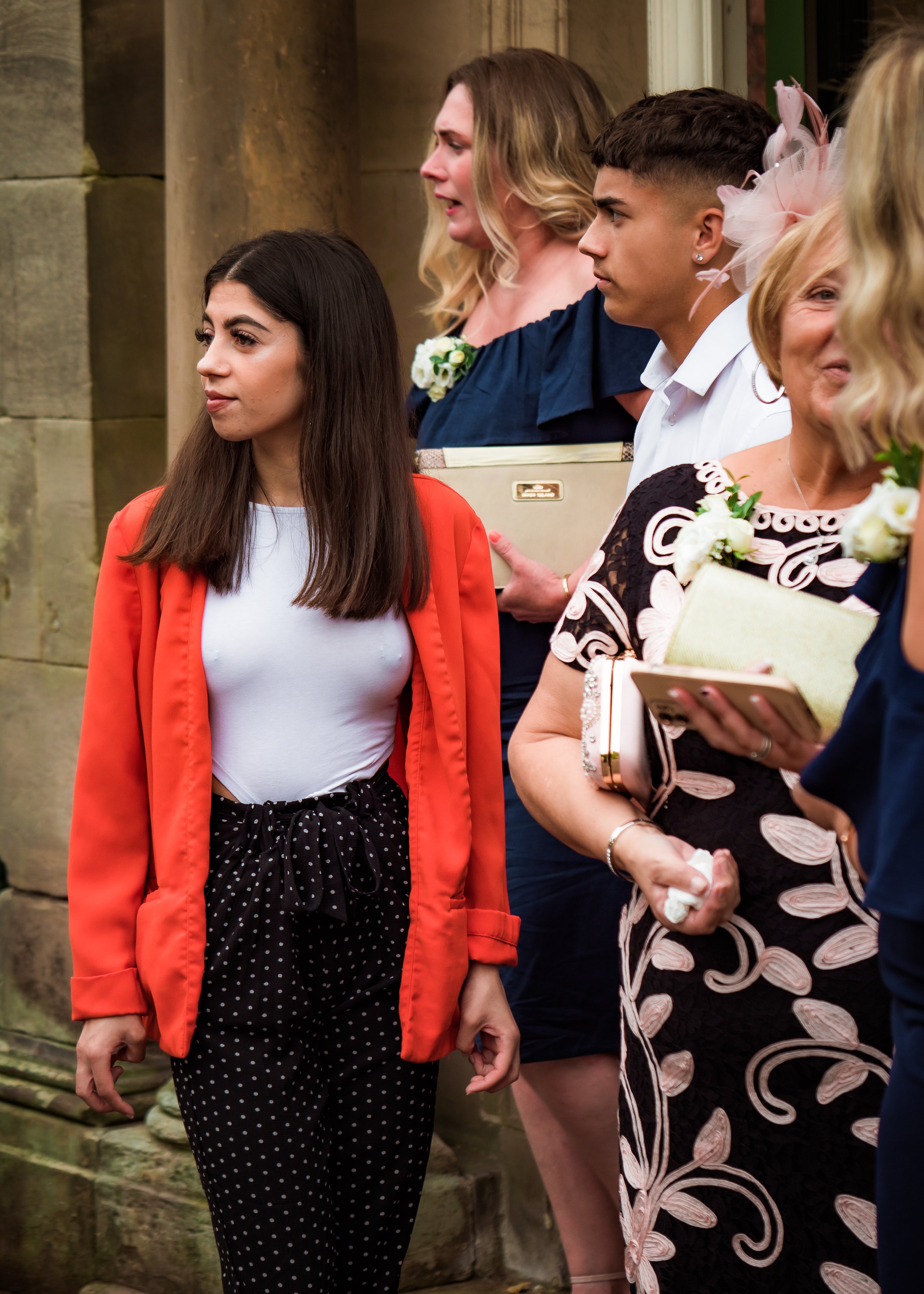 A group of women and a young man standing near a stone wall at an outdoor event, some holding programs and flowers.