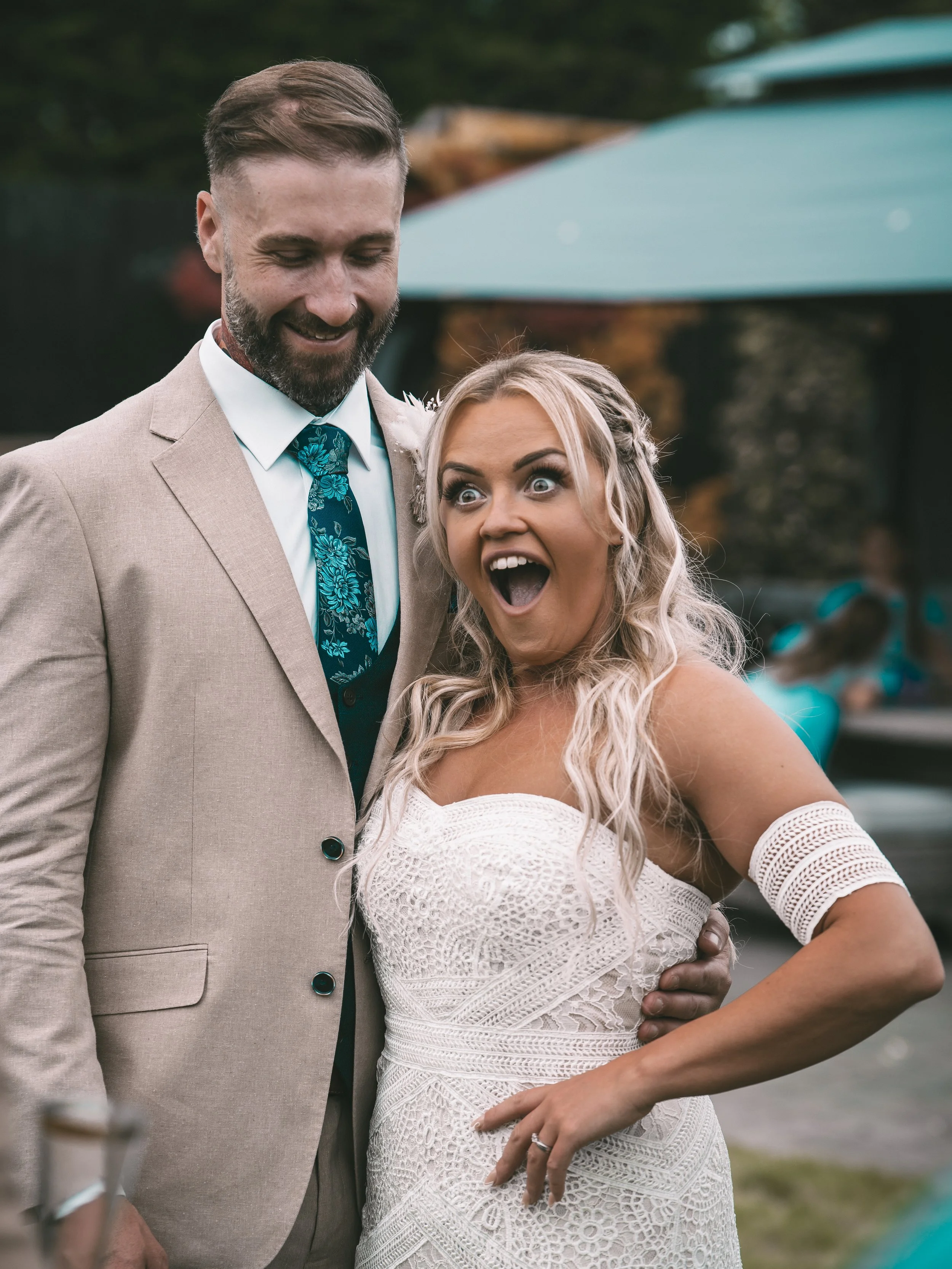 A couple smiling and celebrating outdoors, dressed in wedding attire, with a man in a beige suit and a woman in a white lace dress.