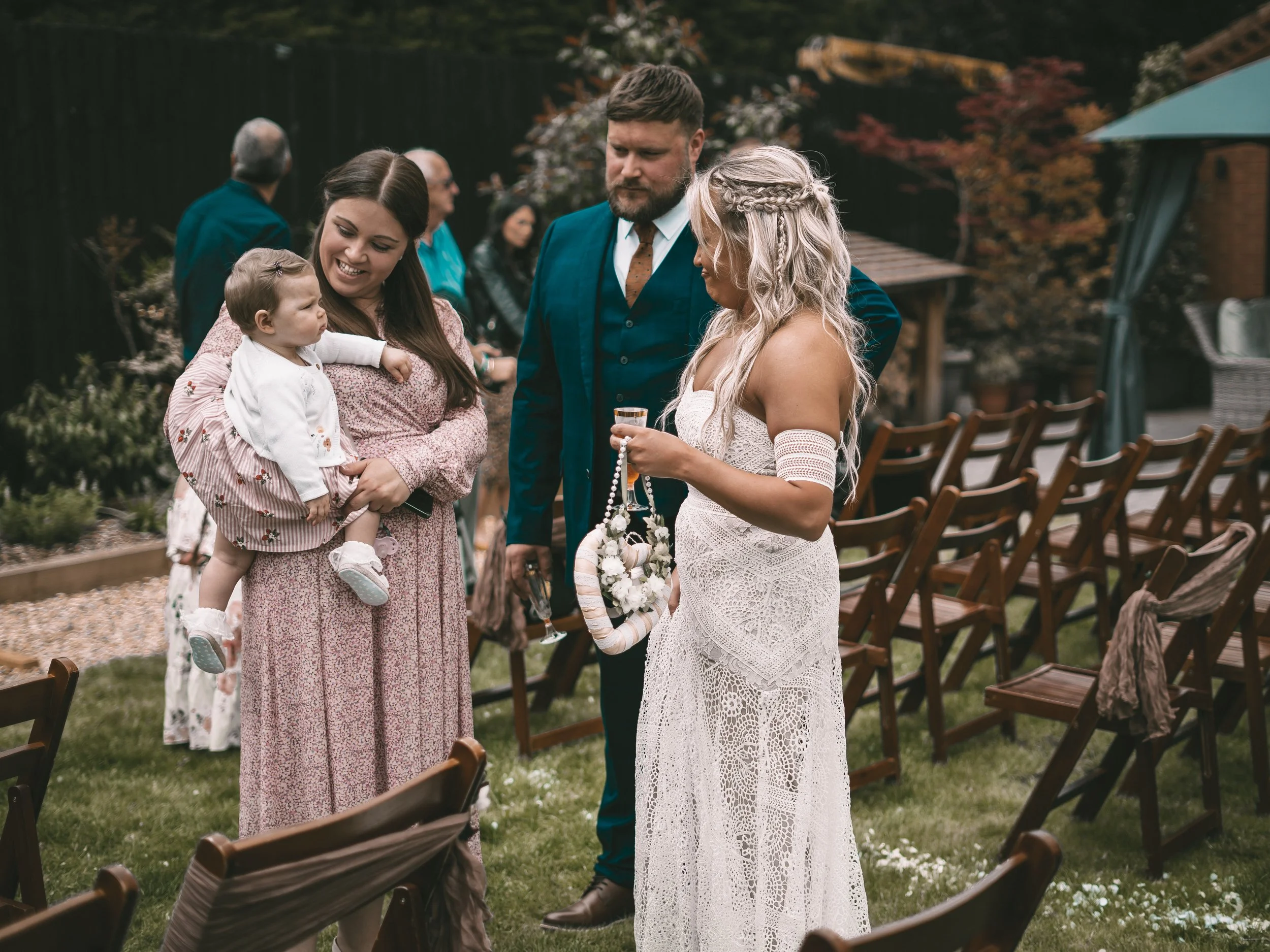 People attending a wedding or outdoor event, with a woman in a white lace dress holding flowers and conversing with a man in a dark suit, while a young girl is held by a woman in a long pink dress, surrounded by chairs and greenery.