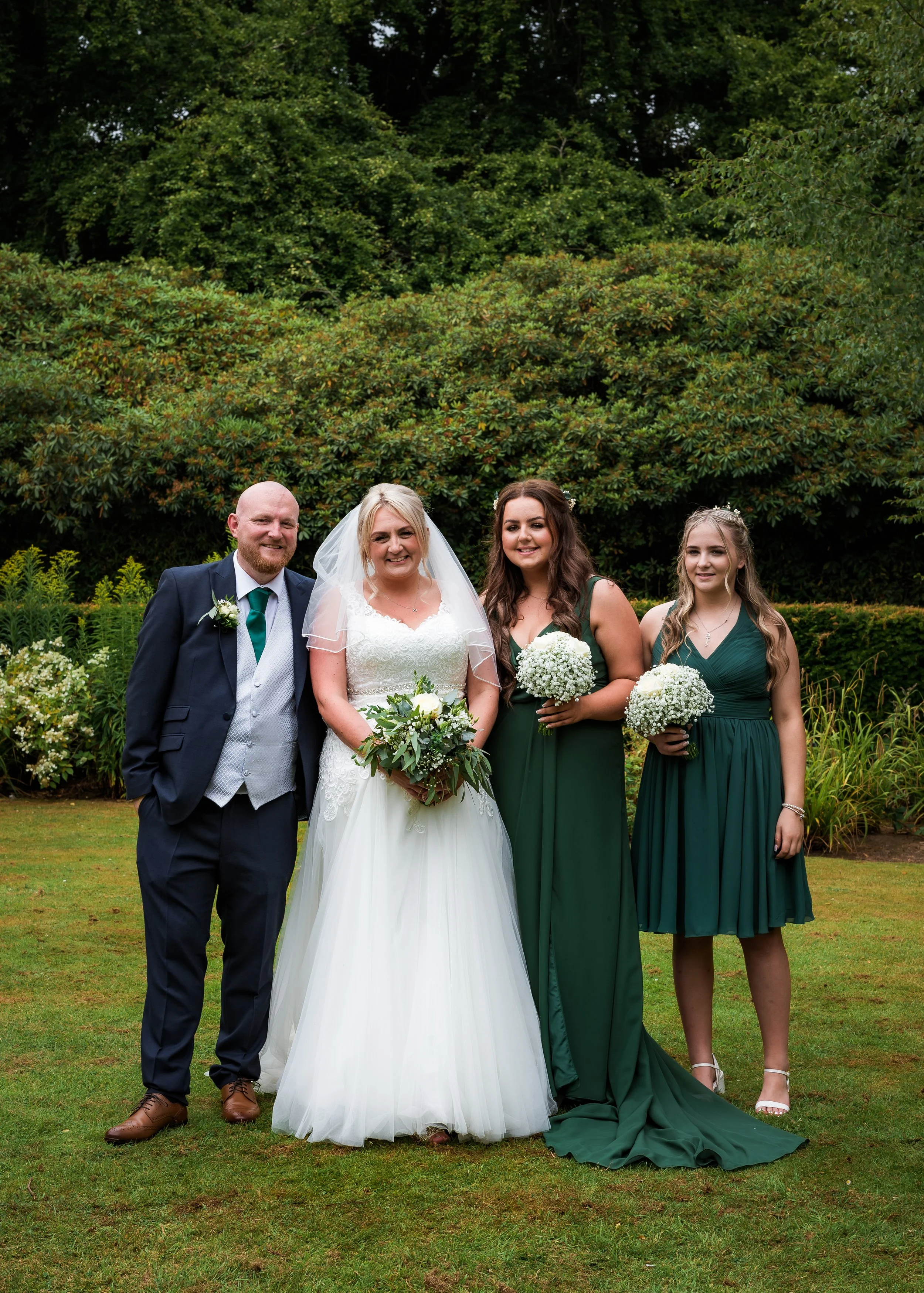 A wedding party consisting of four people: a groom in a navy suit, a bride in a white wedding dress and veil, and two bridesmaids in matching dark green dresses, posing outdoors on a lawn with lush green trees in the background, holding bouquets of w