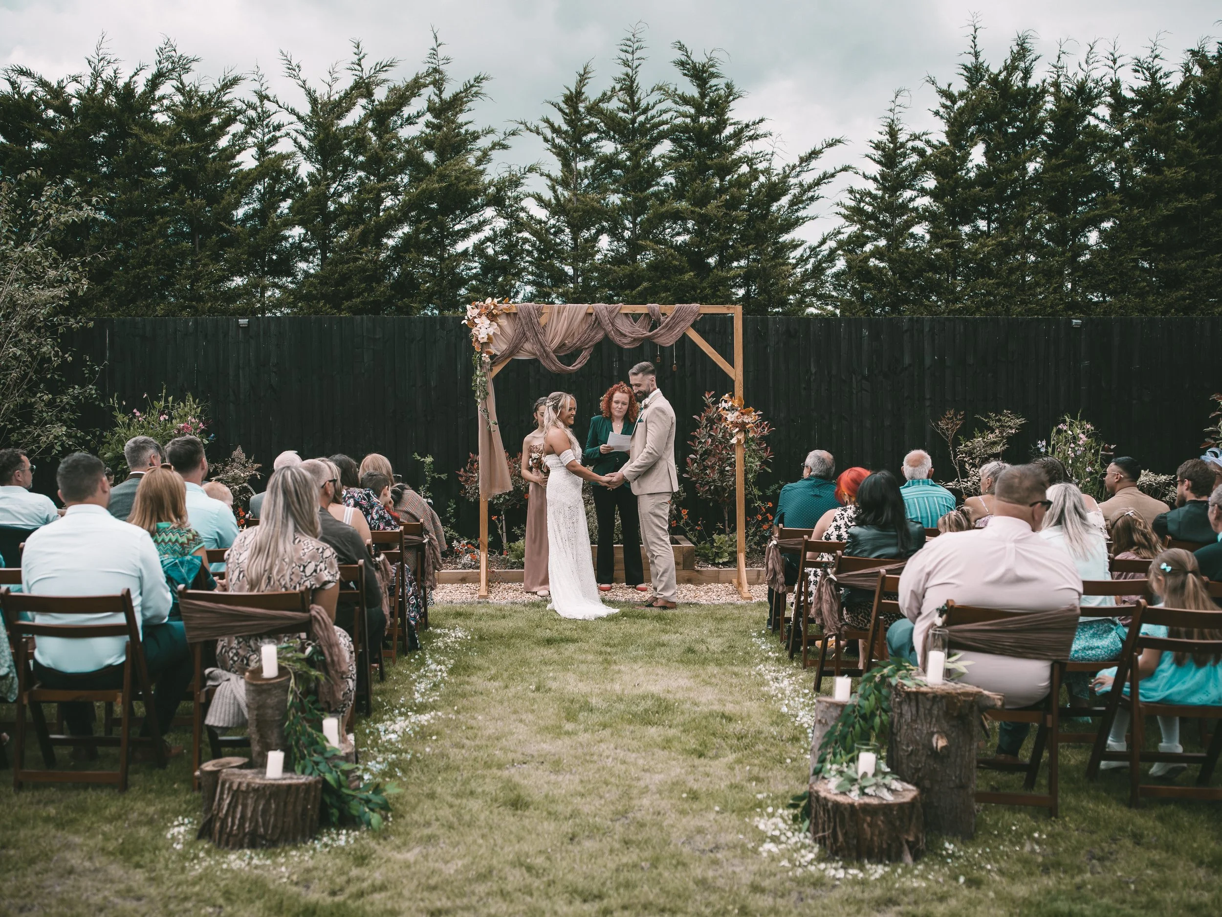Outdoor wedding ceremony with a bride and groom holding hands, standing under a wooden arch decorated with flowers and draped fabric. Guests seated on either side, greenery, and forest trees in the background.