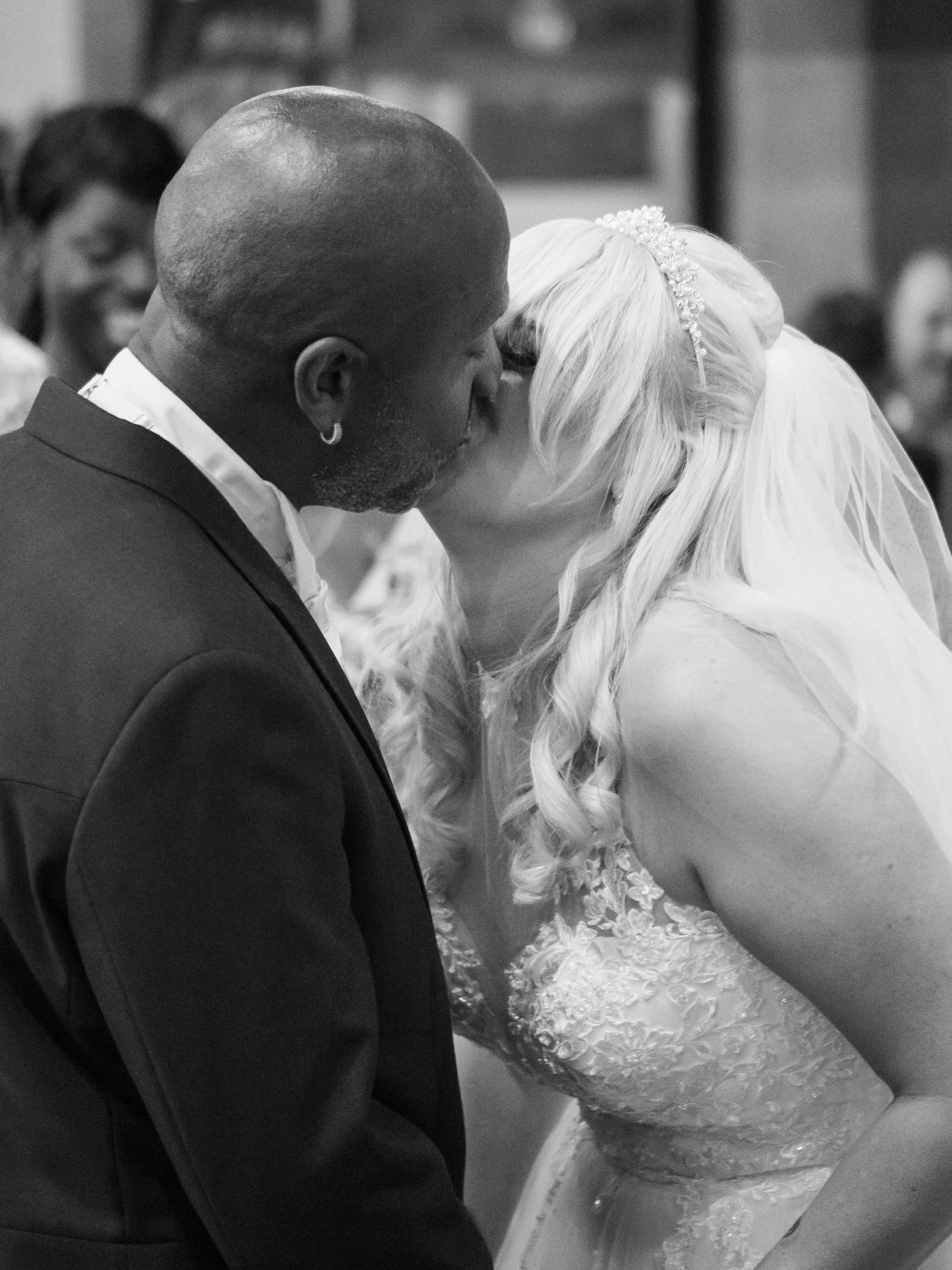 A man and woman kissing during a wedding ceremony, with the man in a suit and the woman in a wedding dress with lace details and a tiara, in a black and white photo.