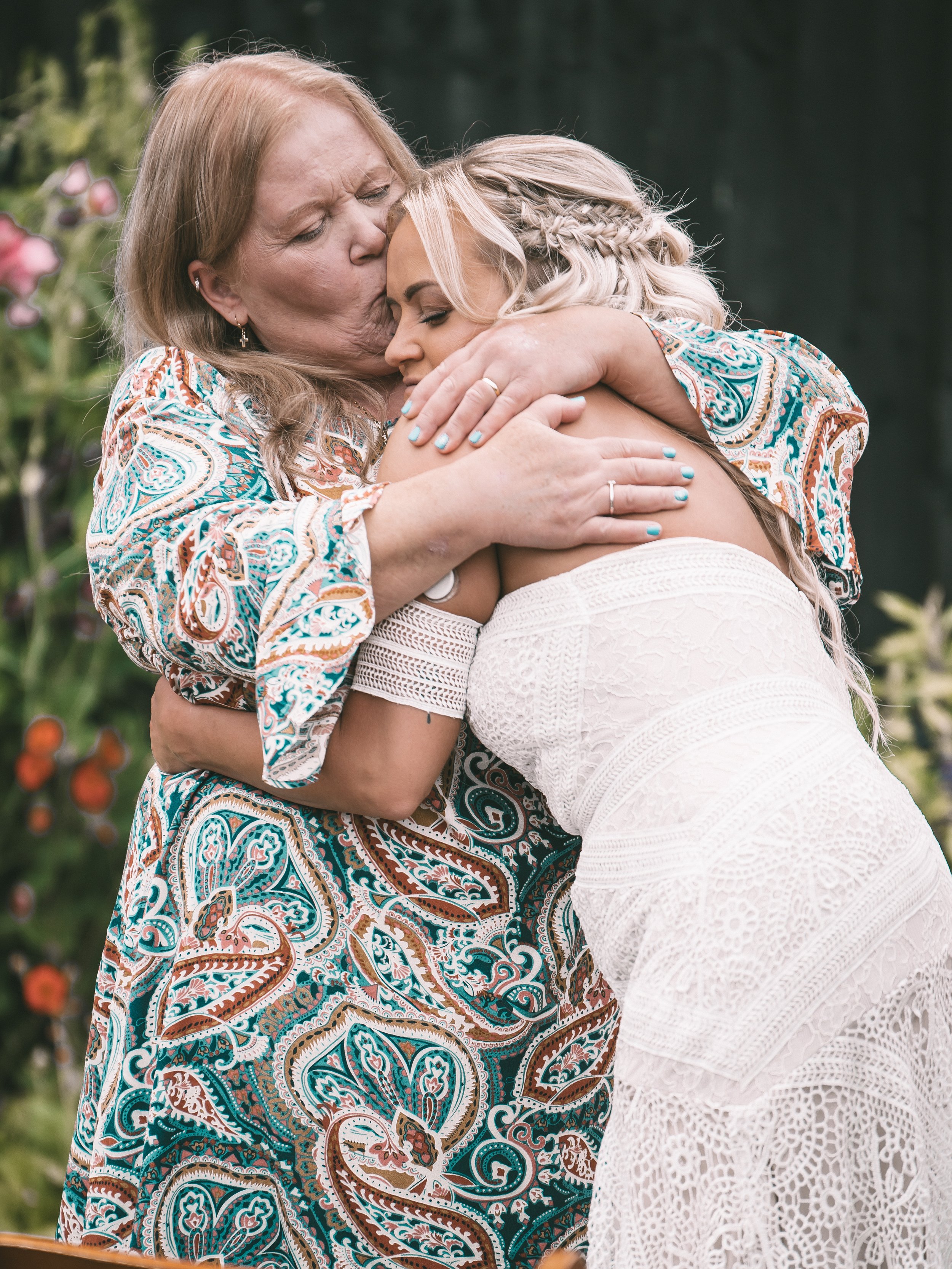 A woman with red hair wearing a patterned dress kisses and hugs a blonde woman wearing a white lace dress in an outdoor garden setting.