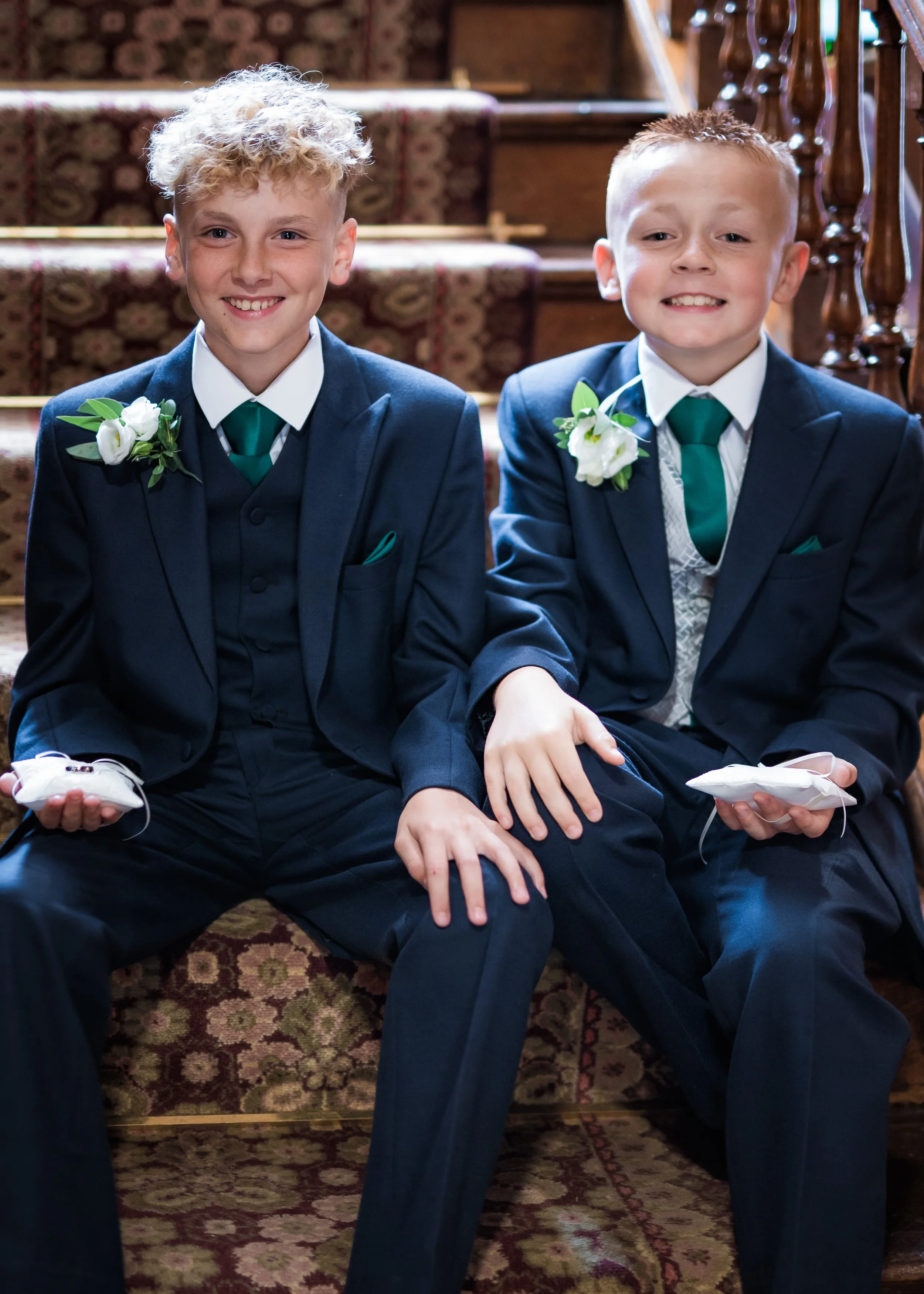 Two young boys dressed in formal dark blue suits with green ties and white shirts, sitting on a staircase with floral carpet, smiling at the camera, each holding a white face mask.