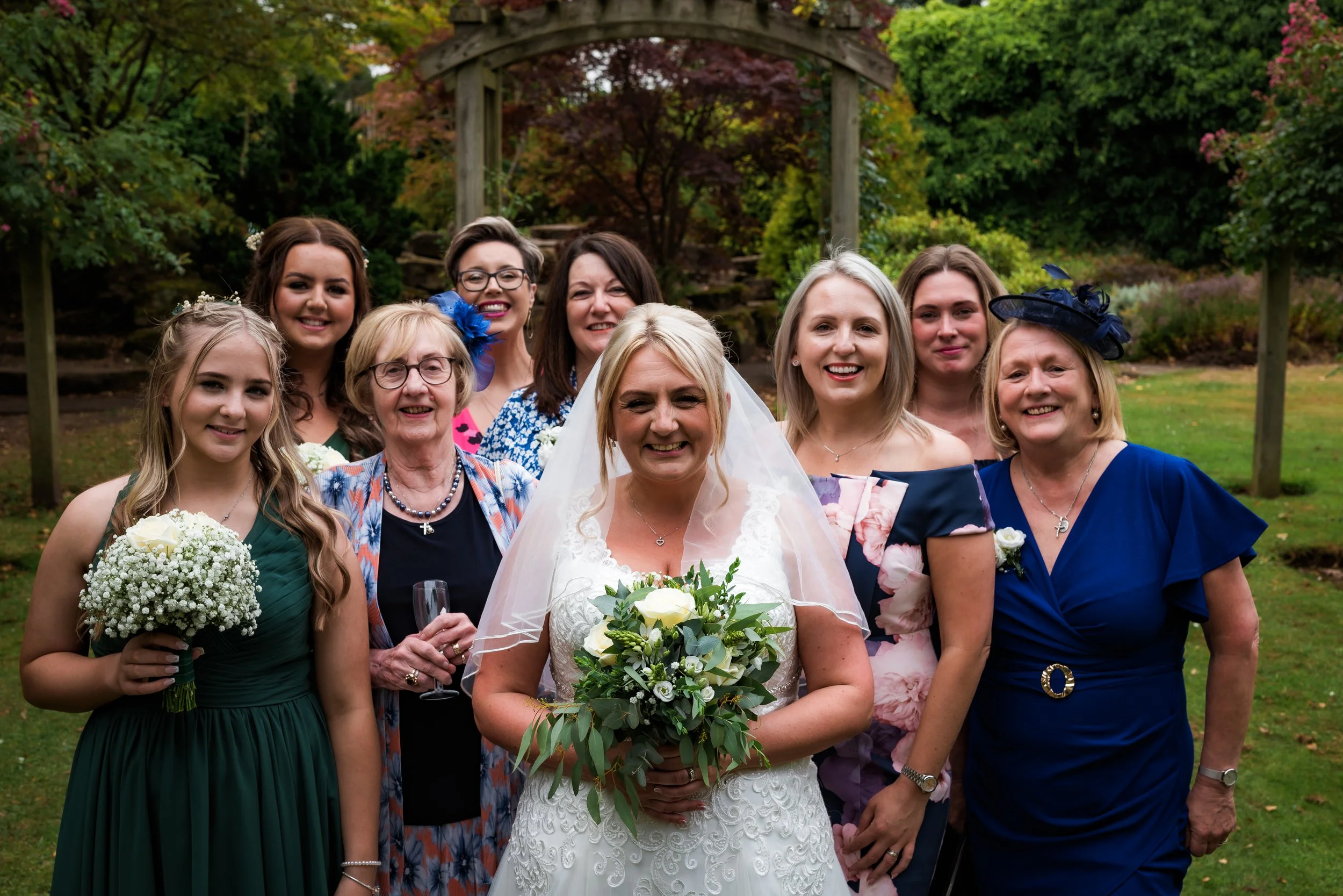 A group of women, including a bride holding a bouquet, posing outdoors in front of a garden with autumn foliage and a wooden arch.
