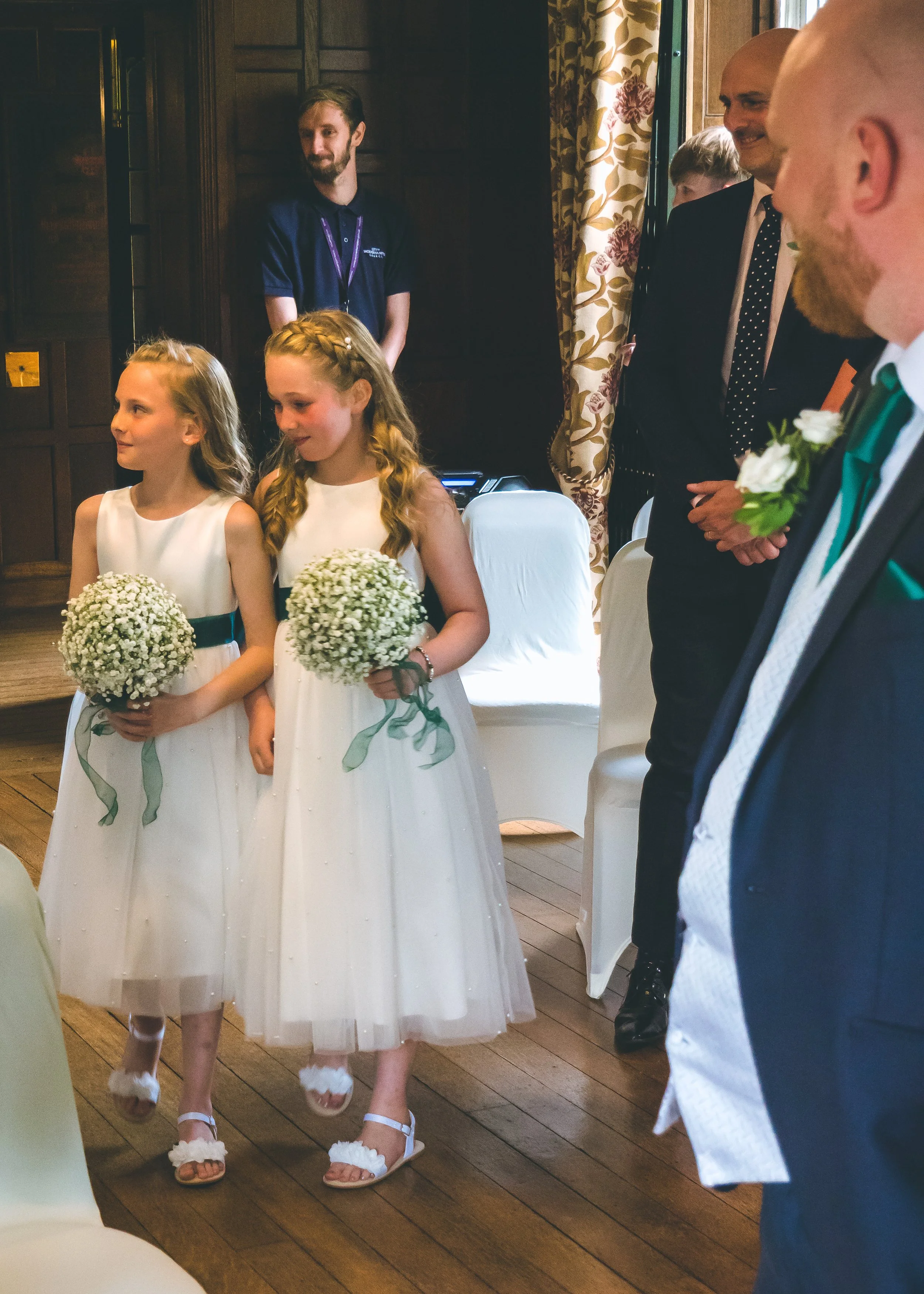Two young girls in white dresses holding bouquets of white flowers standing in front of seated guests during a wedding ceremony in a cozy, wood-paneled room with floral curtains.
