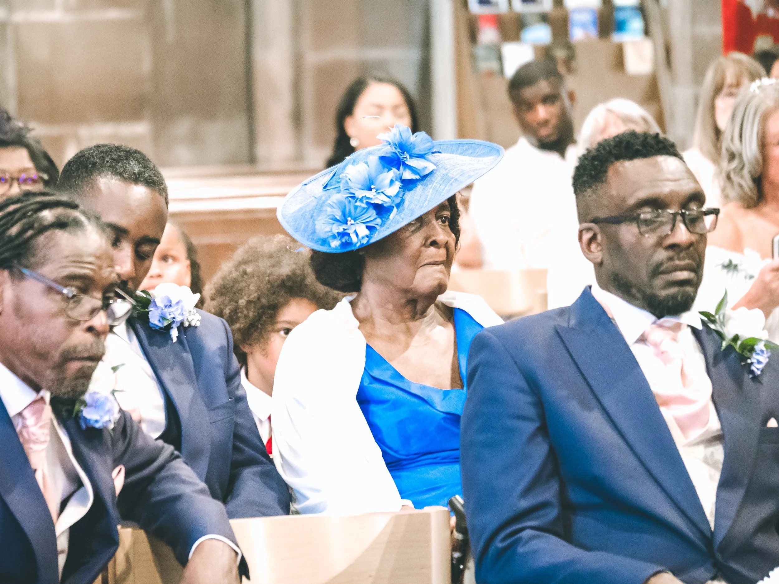 A group of people attending a formal event, sitting in rows. A woman in the center wears a large blue hat with flowers, and a woman to her right is dressed in a blue outfit. The audience members are dressed in suits and dresses, and some have boutonn