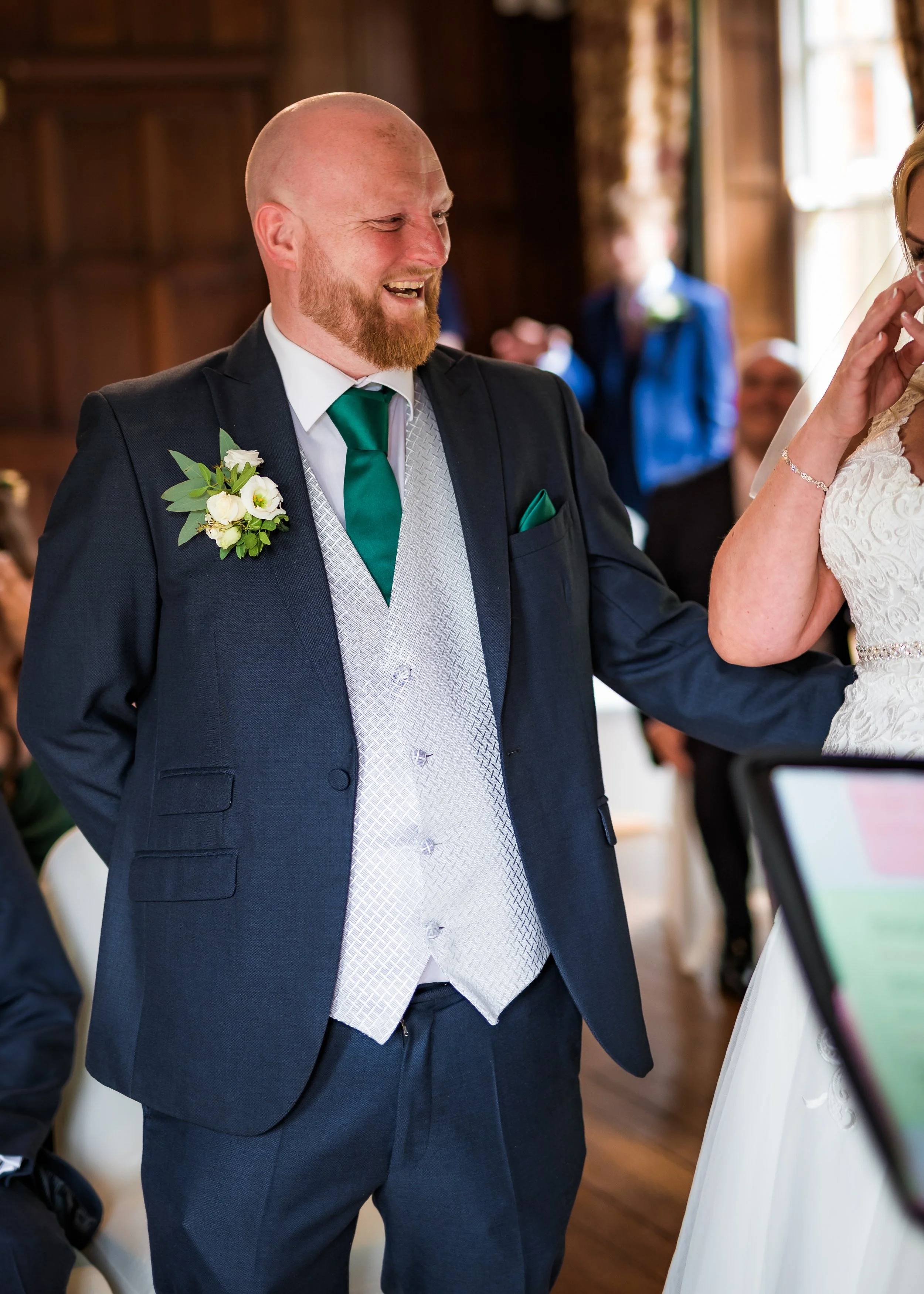 A man in a suit with a boutonniere, smiling during a wedding ceremony, standing close to a woman in a white dress.