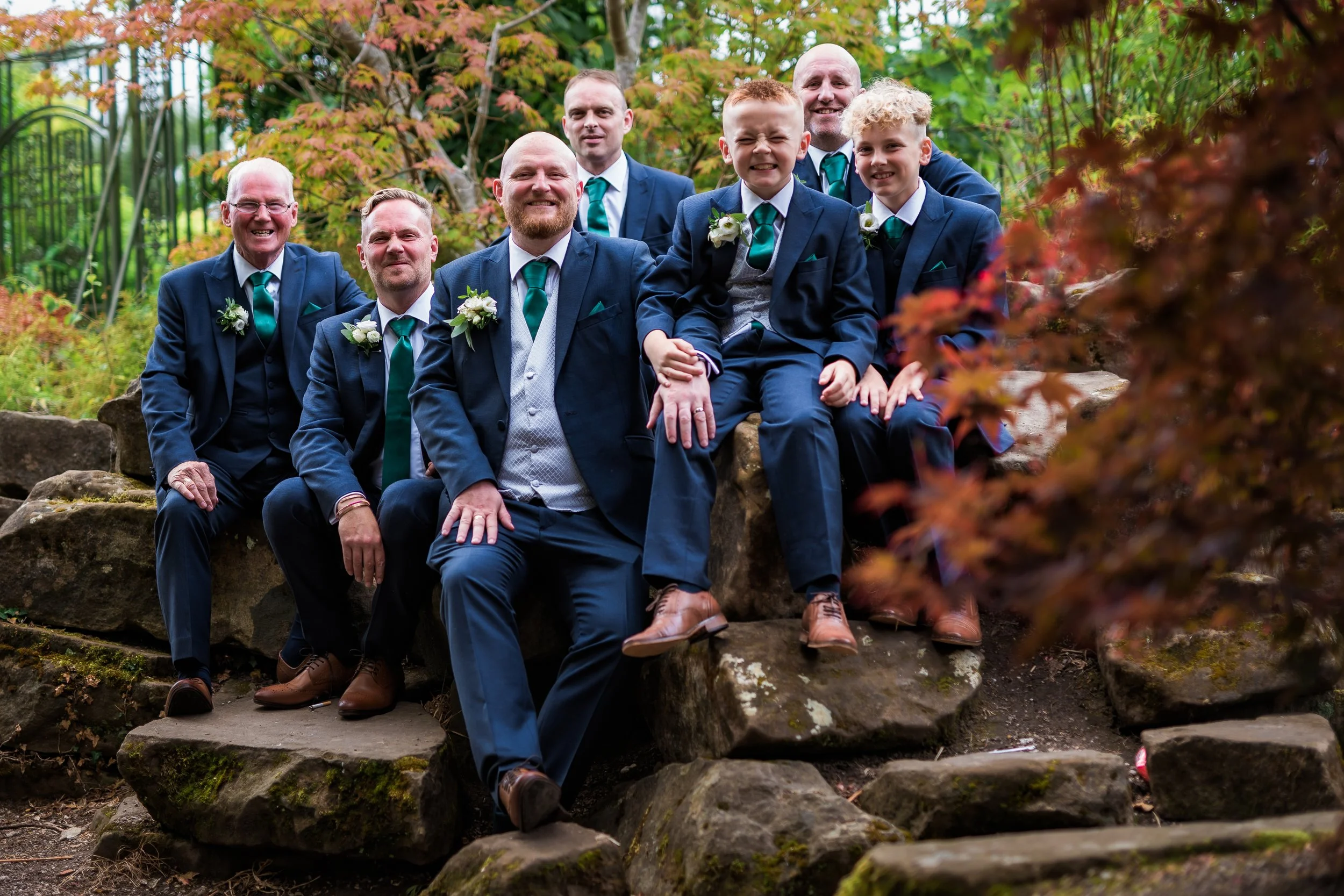 A group of eight men in suits with teal ties and white boutonnieres sitting and standing on rocks outdoors. The background features autumn-colored trees and a metal fence.
