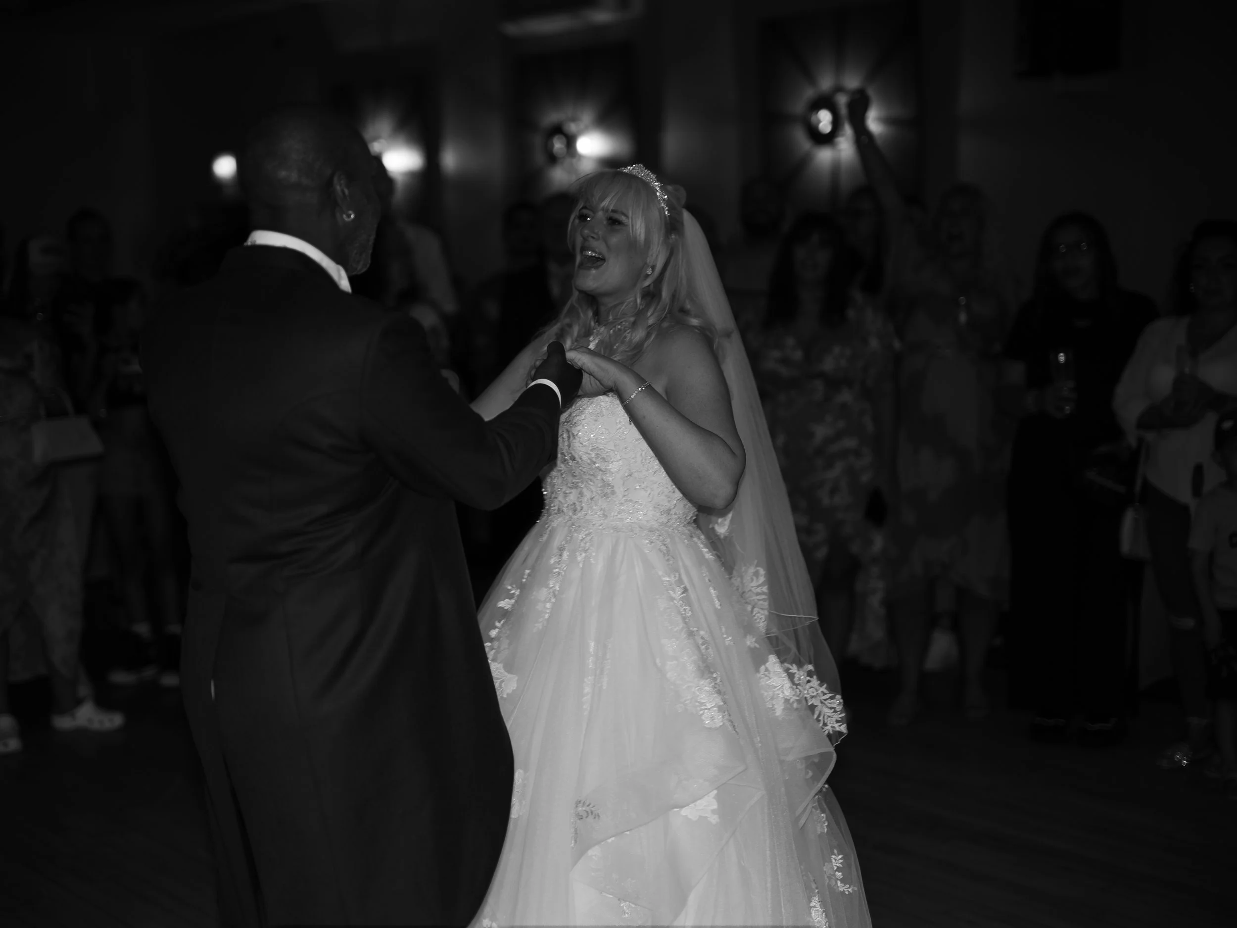 A bride and groom dancing at their wedding reception with guests observing in the background, black and white photo.