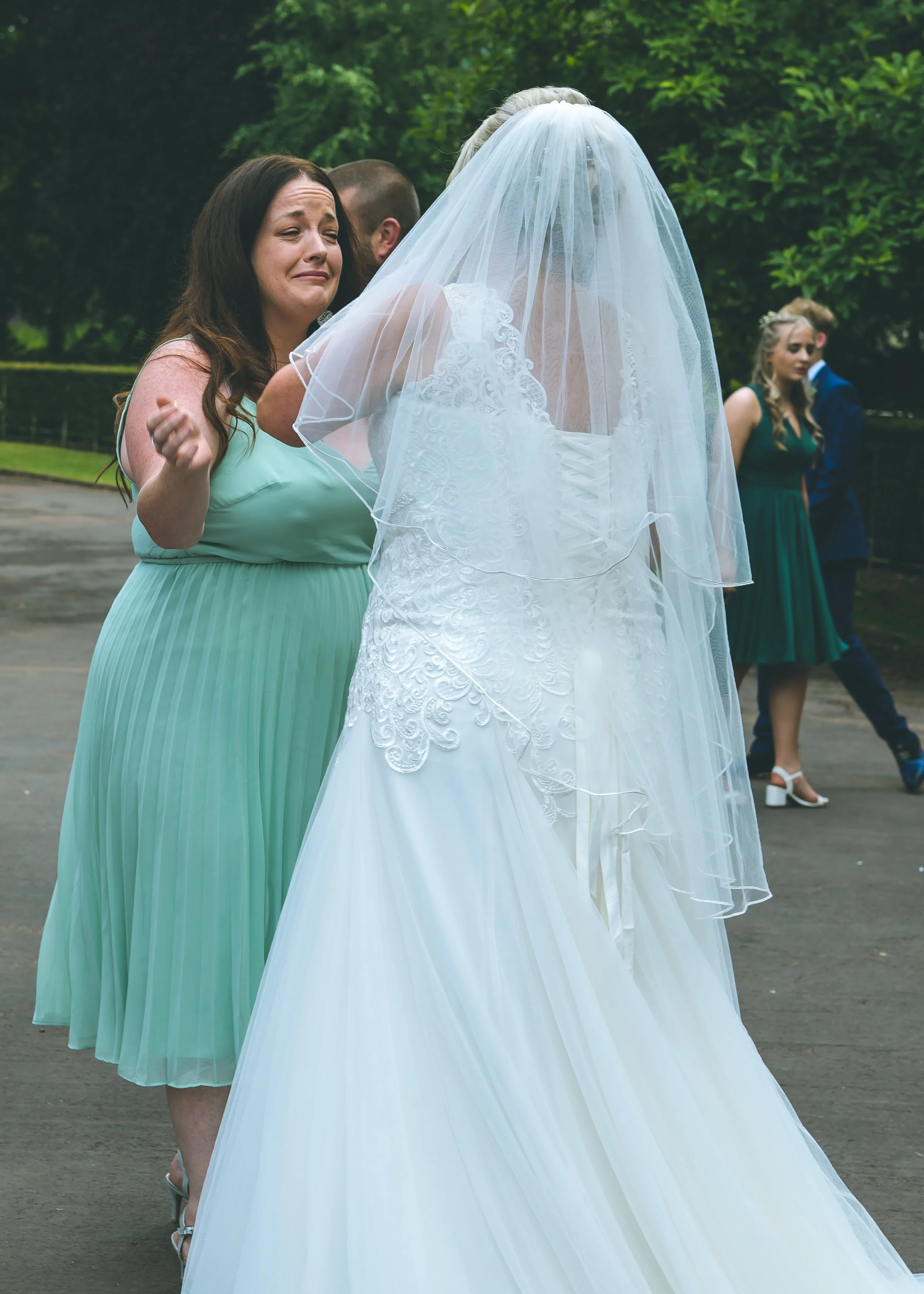 A woman crying and emotional during a wedding ceremony, standing in front of a bride in a white wedding gown with a long veil, outdoors with green trees in the background. Other guests in formal attire are visible in the background.