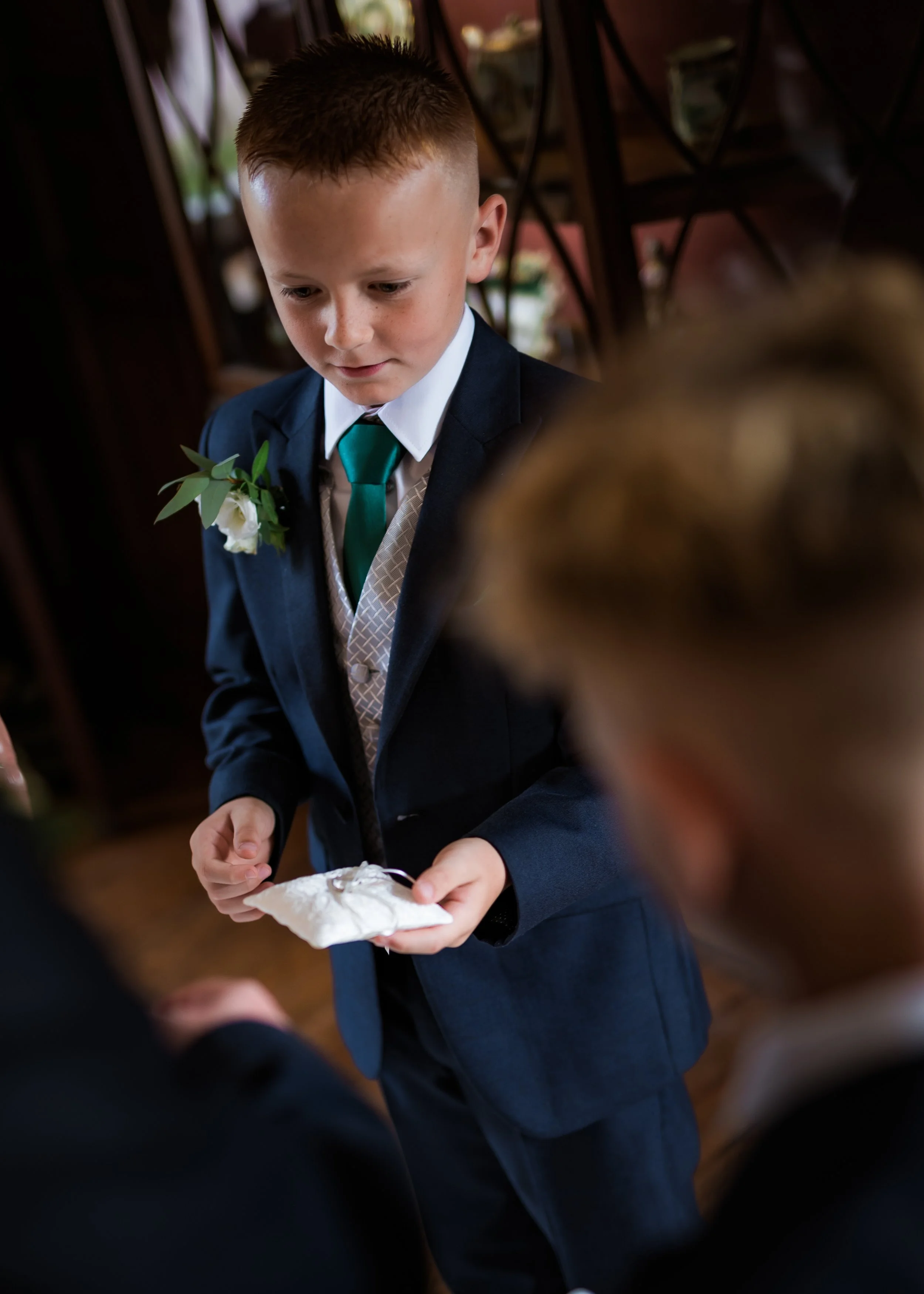 A young boy dressed in a navy suit with a white shirt and green tie, holding a white pillow, possibly during a wedding ceremony, with floral decorations in the background.