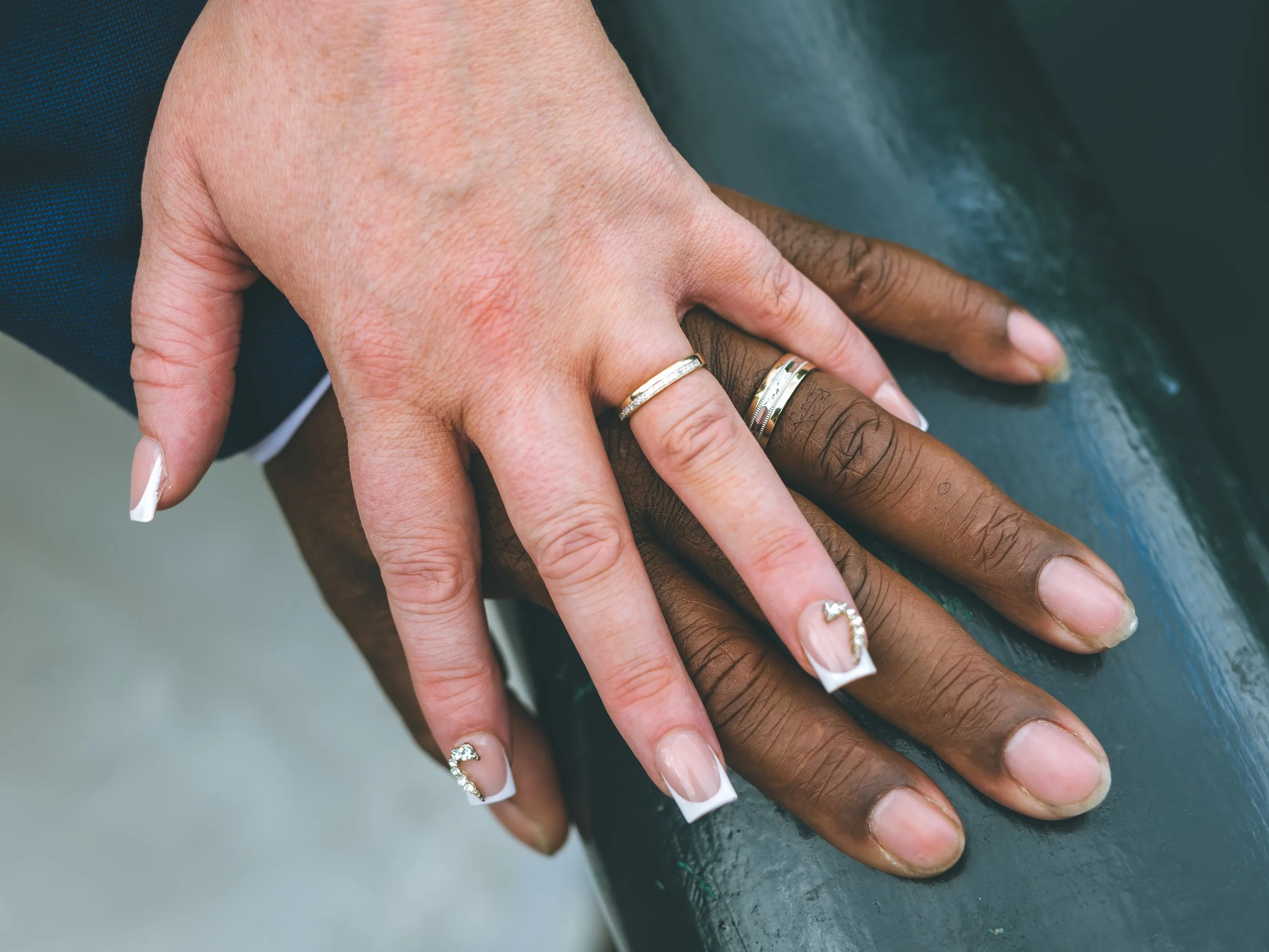 Close-up of two hands, one with a lighter skin tone and the other with a darker skin tone, resting on a surface. Both hands are wearing wedding rings.
