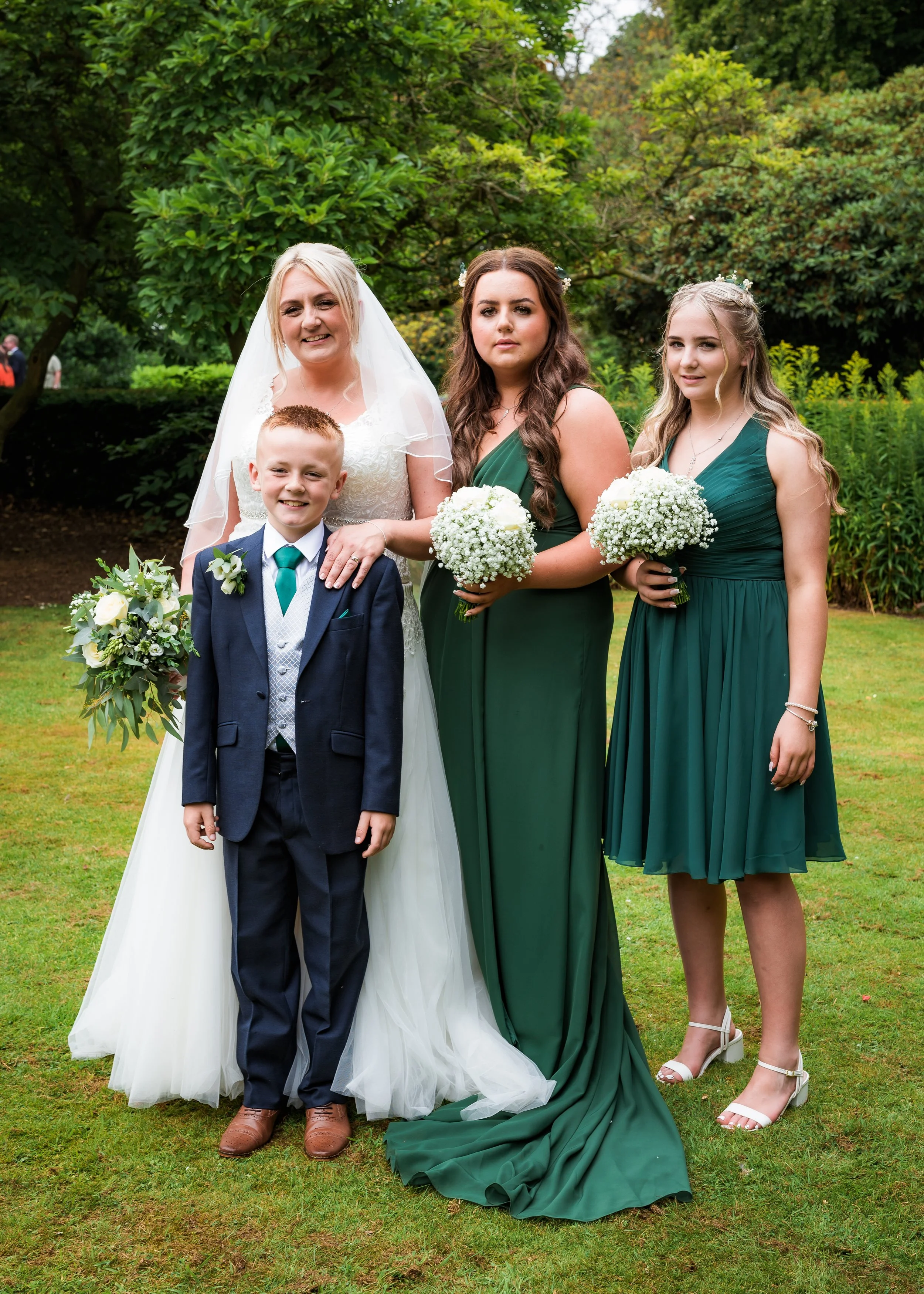Group of four people at a wedding outdoors, including a bride in a white wedding dress and veil, a young boy in a suit holding a bouquet, and two young women in green dresses holding bouquets, standing on grass with green trees behind them.