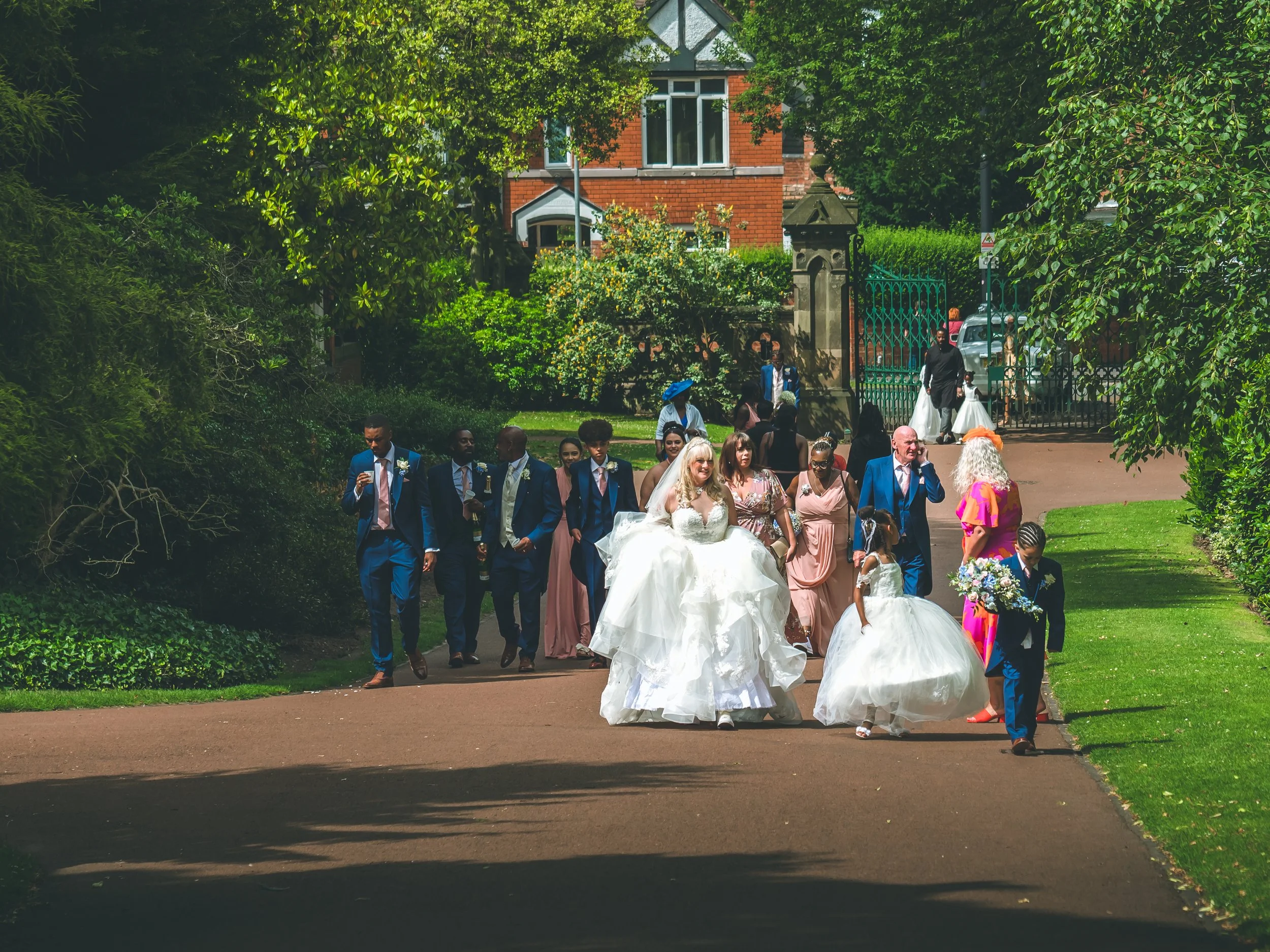 A group of wedding guests, including a bride in a white gown, walking along a pathway outside a red-brick house covered by large trees, with some women wearing pastel dresses and men in suits, as part of a wedding celebration.