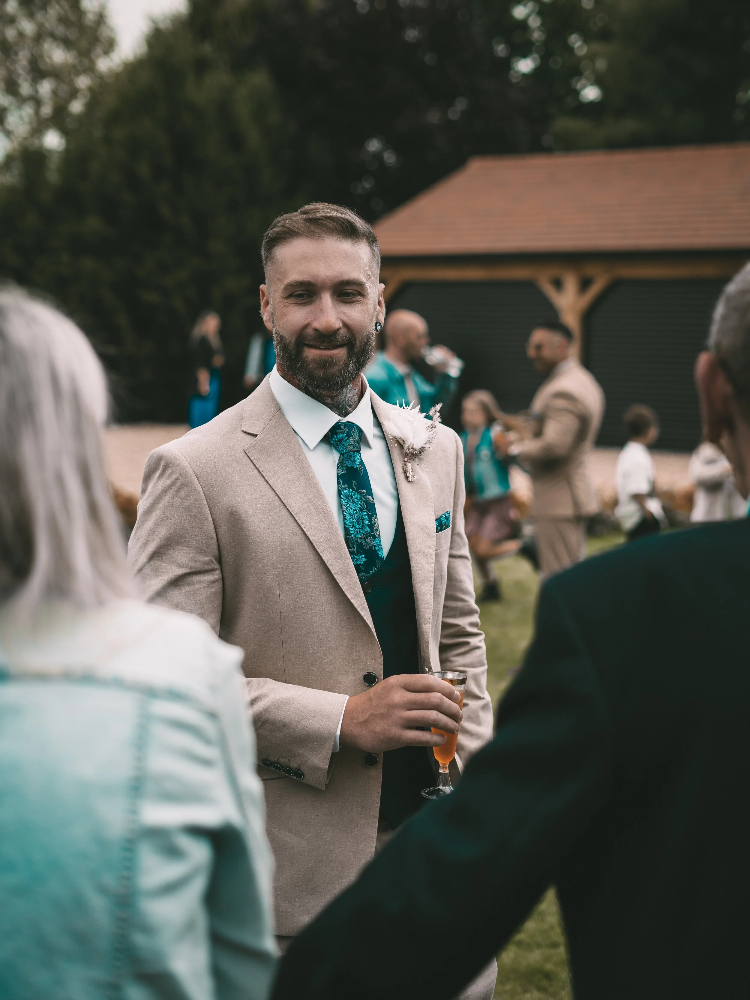 A man with a beard and tattoos, wearing a beige suit, white shirt, and teal tie with floral pattern, holding a glass of champagne at an outdoor gathering. In the background, other guests are mingling and a black barn with a wooden roof is visible.